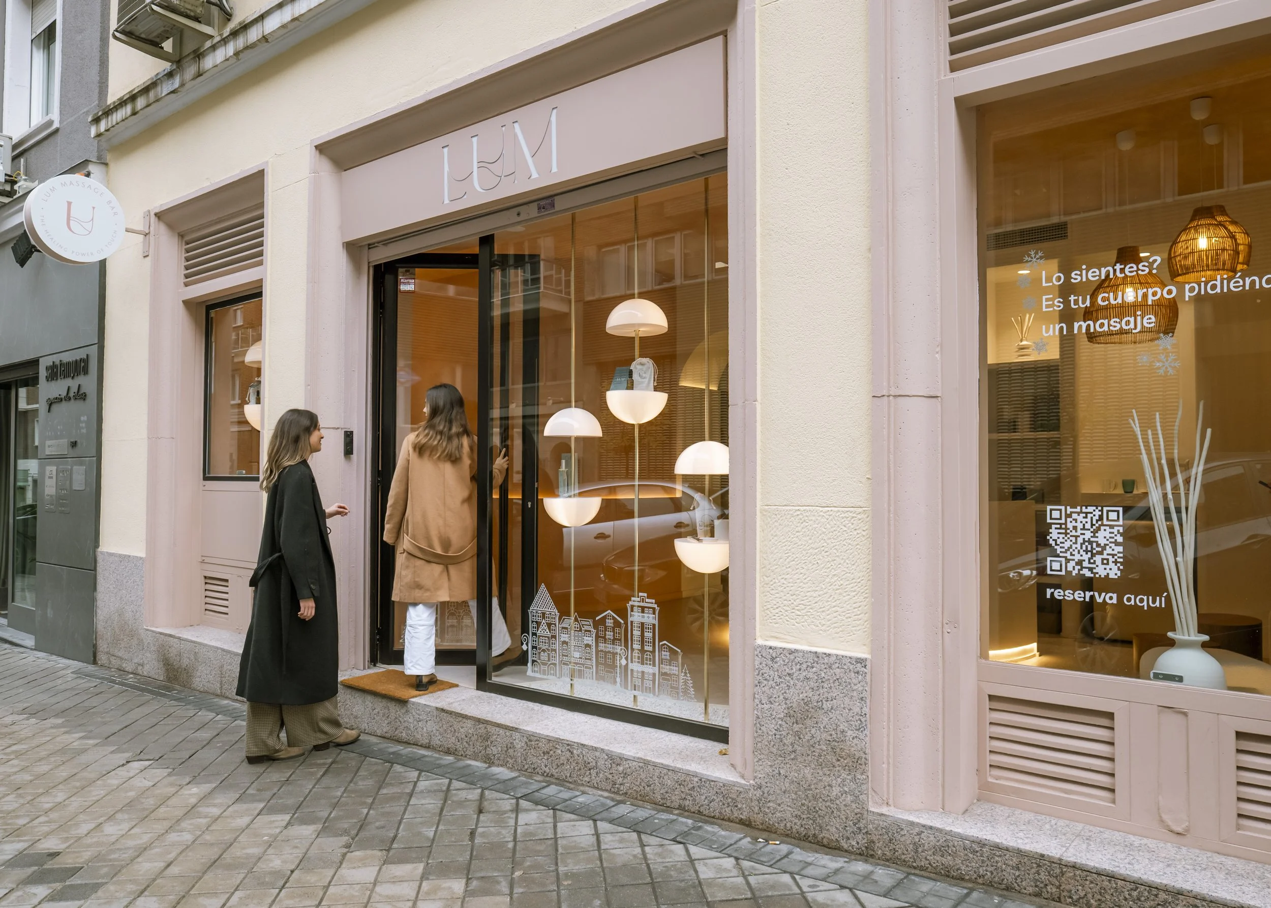 Dos mujeres entrando a un spa o salón de masajes en una calle peatonal. La fachada tiene pantallas con decoraciones modernas y letras que dicen 'LUM'.