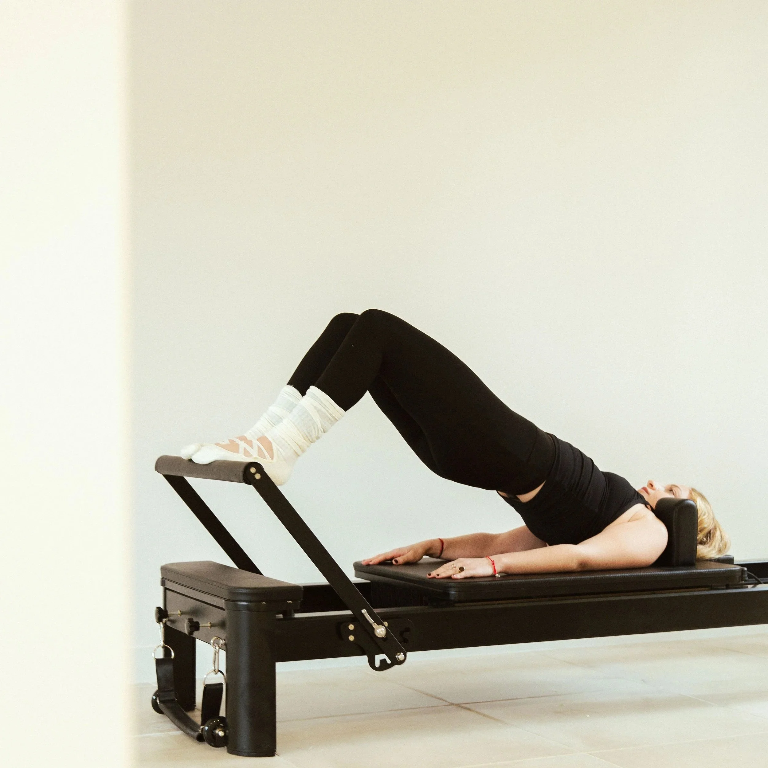 Femme allongée sur un appareil de Pilates, effectuant un exercice avec les jambes sur une plateforme inclinée, portant des vêtements noirs et chaussettes blanches.