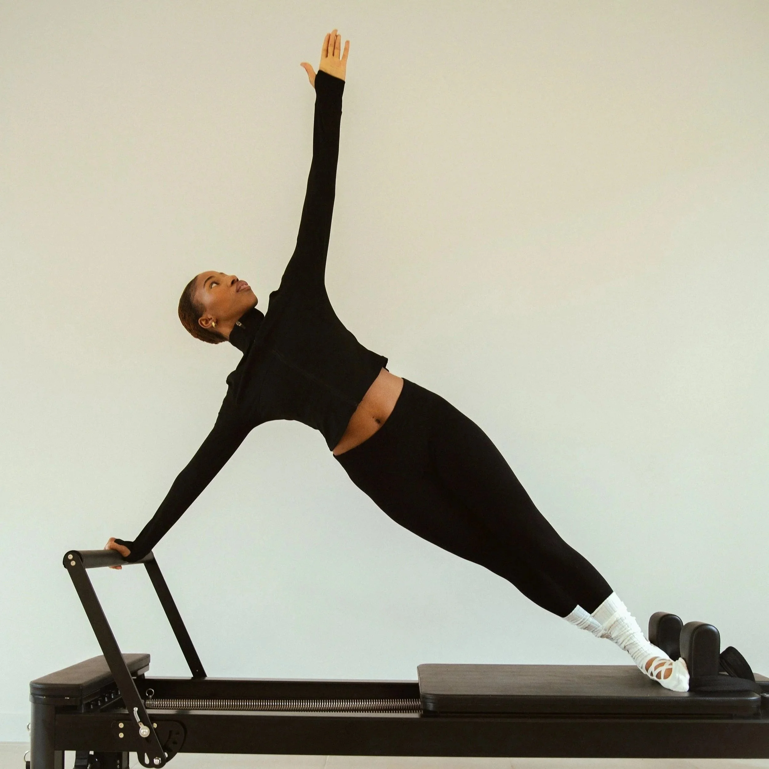 Femme faisant une pose de yoga sur un reformer Pilates, en appui sur une main et les jambes tendues, portant des vêtements de sport noirs et des chaussettes blanches, regardant vers le haut.