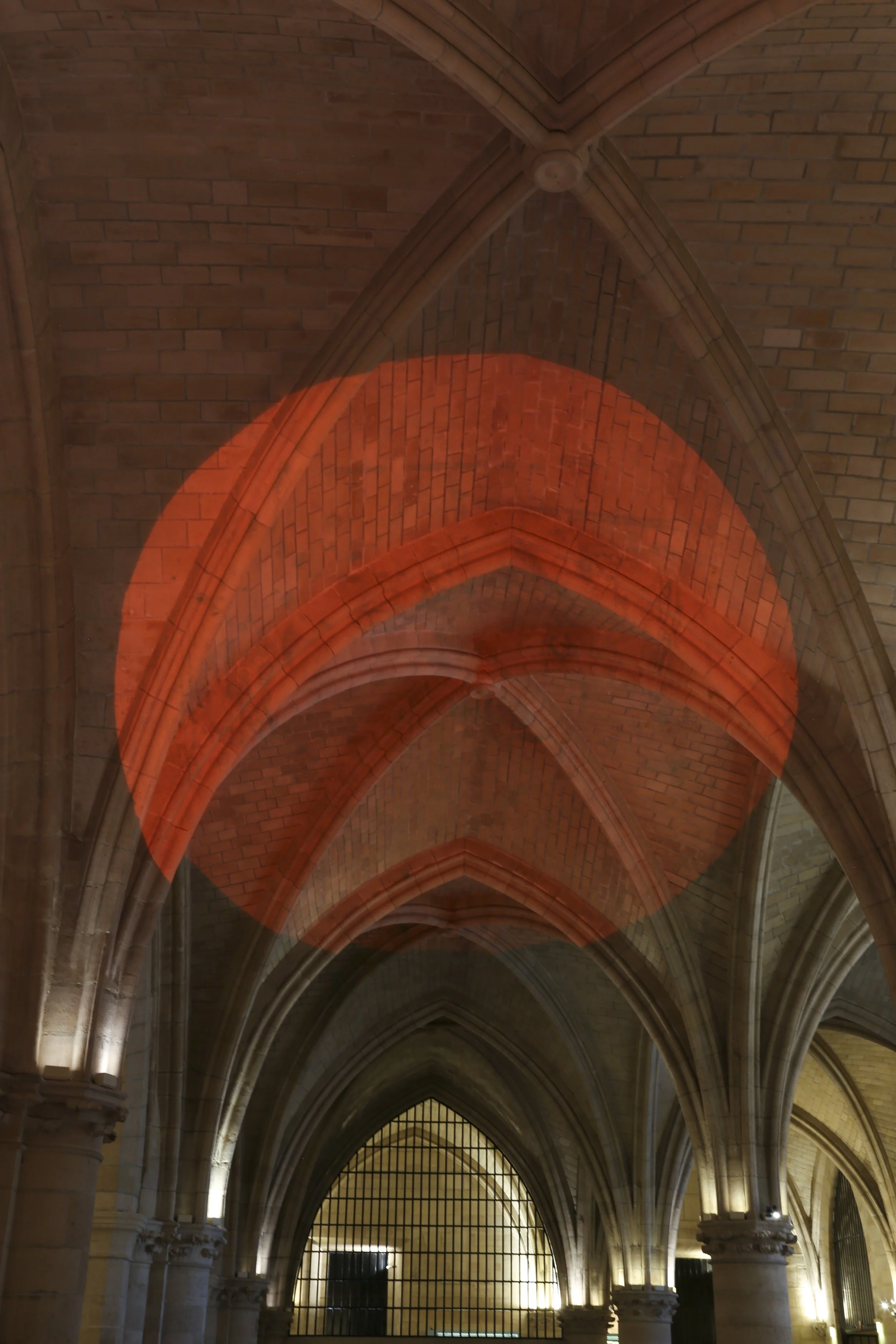 Un plafond voûté en pierre avec des arches gothiques, éclairé par des lumières situées à la base des piliers, avec une lumière rouge projetée sur une partie du plafond.