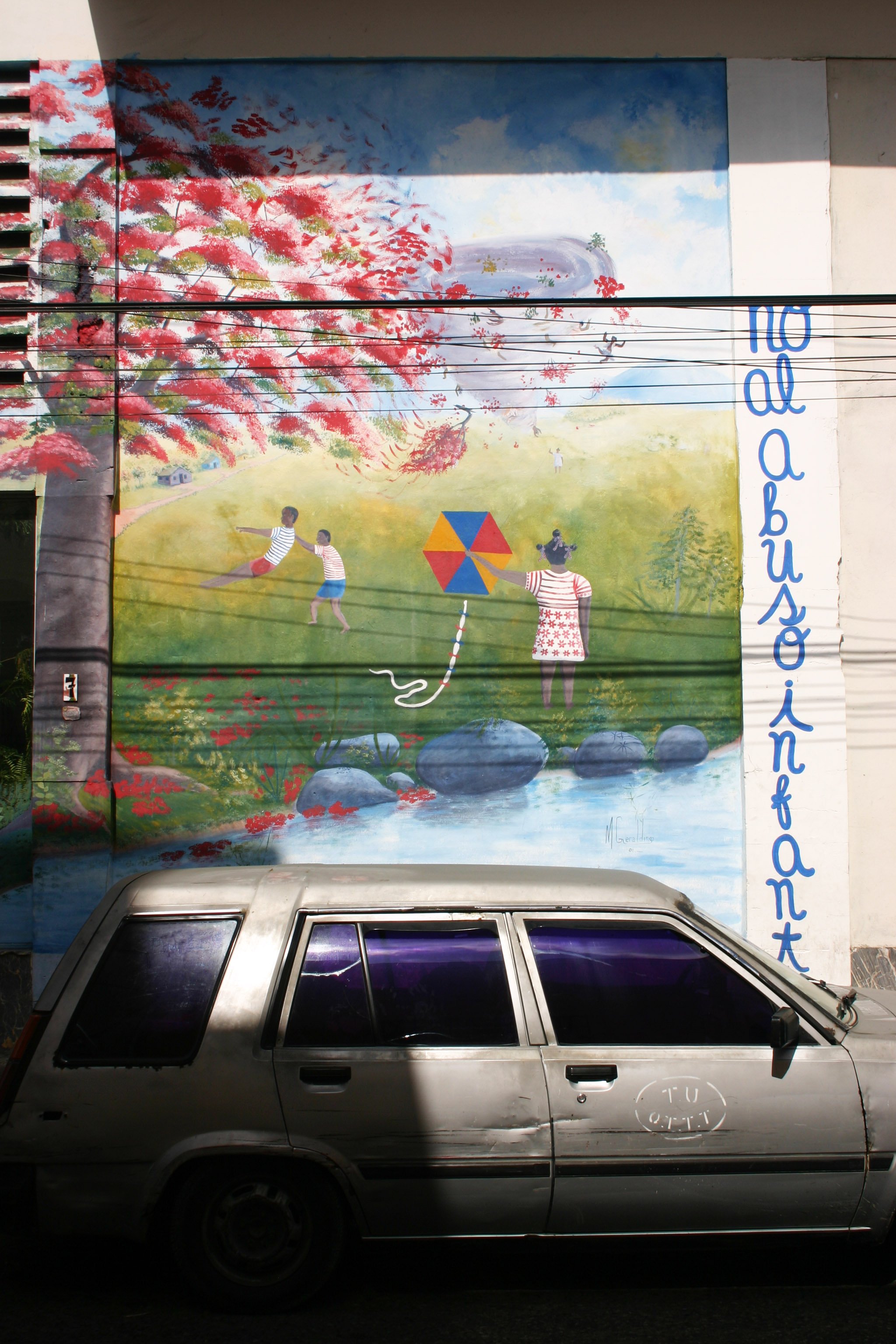 Peinture murale représentant des enfants jouant dans un champ, avec un arbre en fleurs, un cerf-volant et un ciel bleu, à côté d'une voiture argentée stationnée au premier plan.