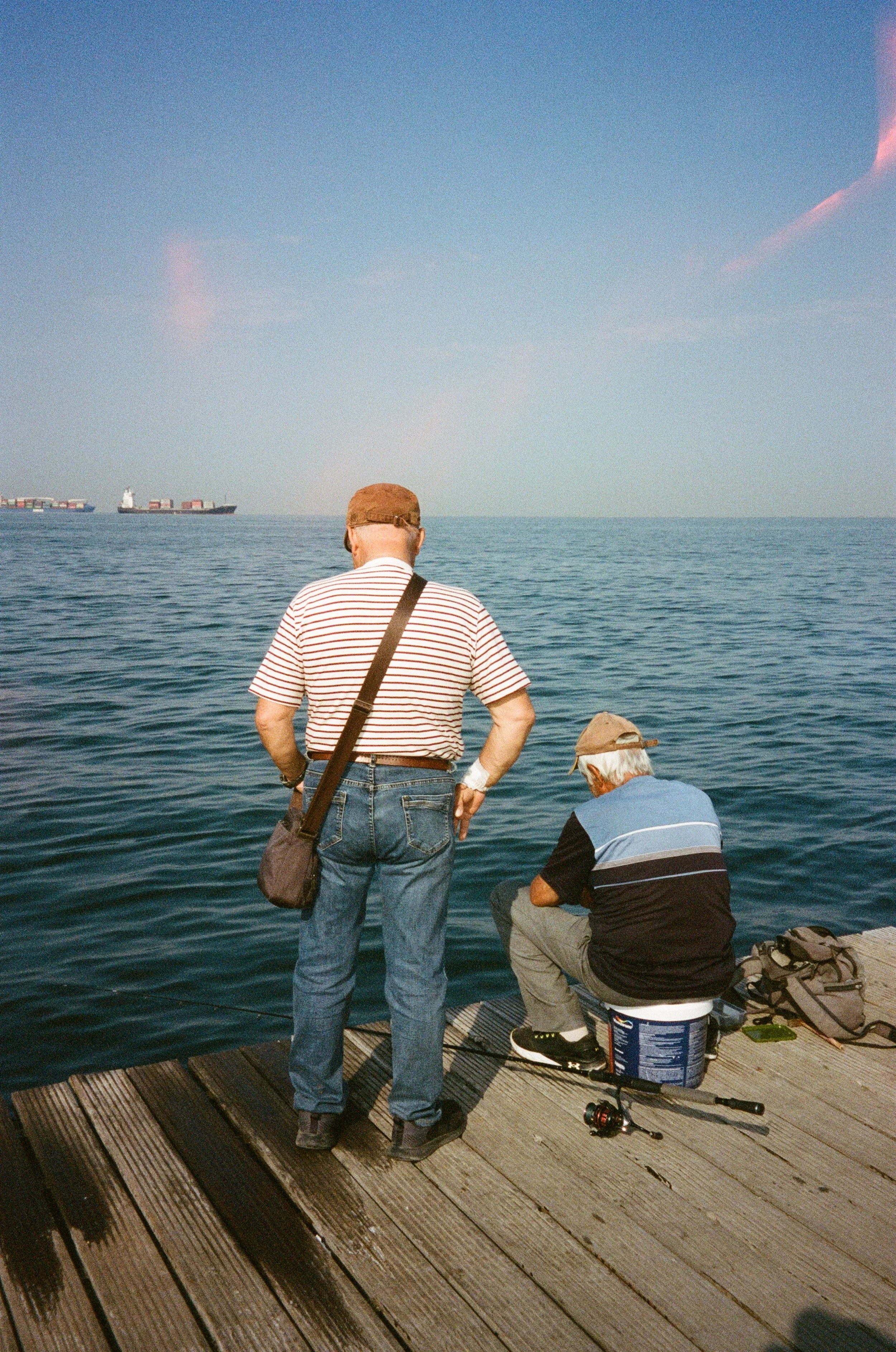 Deux hommes pêcheurs sur un quai en bois en regardant la mer, un avec un rayé blanc et rouge, l'autre en noir et bleu, avec un bateau cargo en arrière-plan.