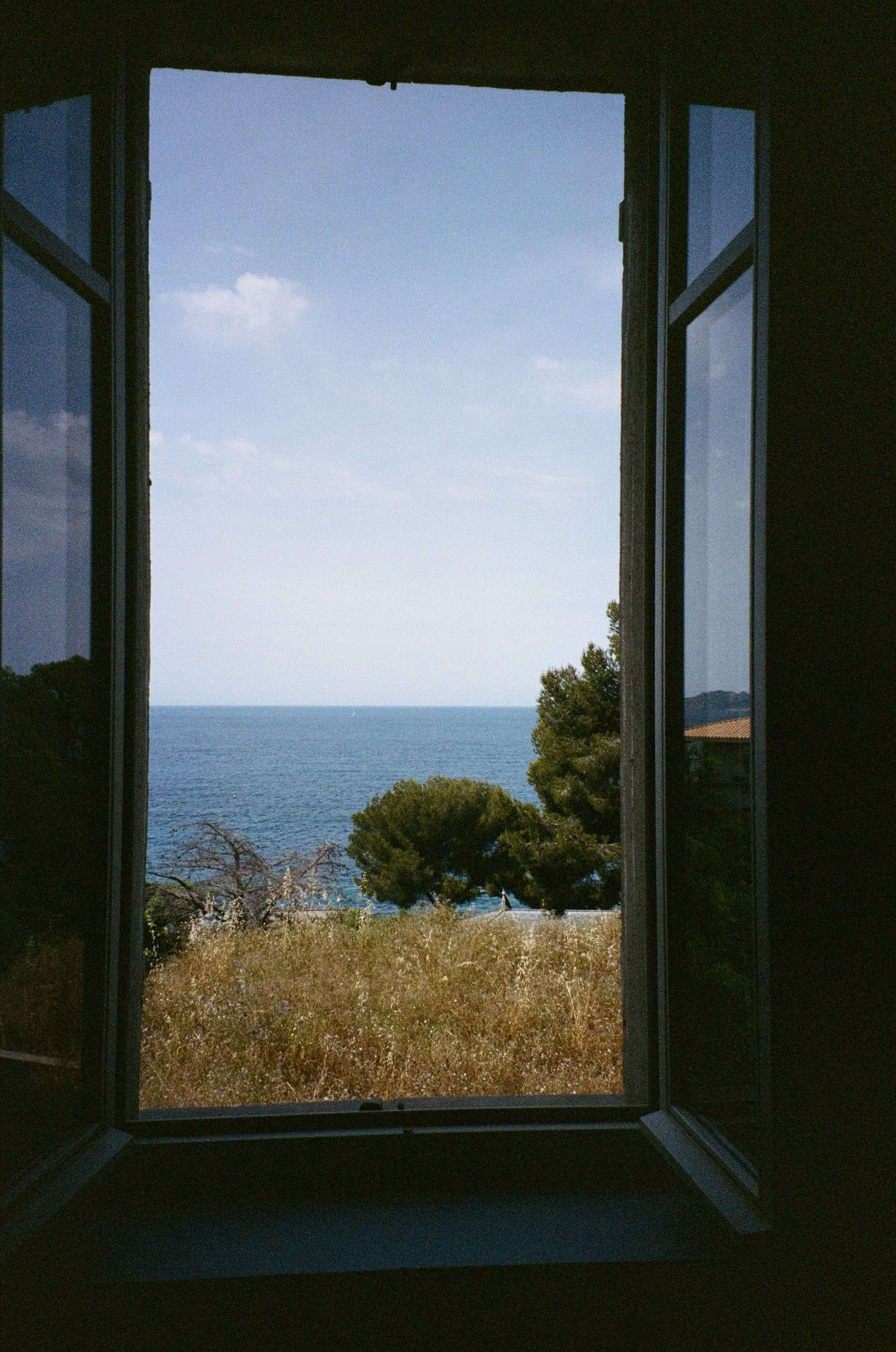 Vue de la mer à travers une fenêtre ouverte, avec des arbres et un ciel bleu clair.