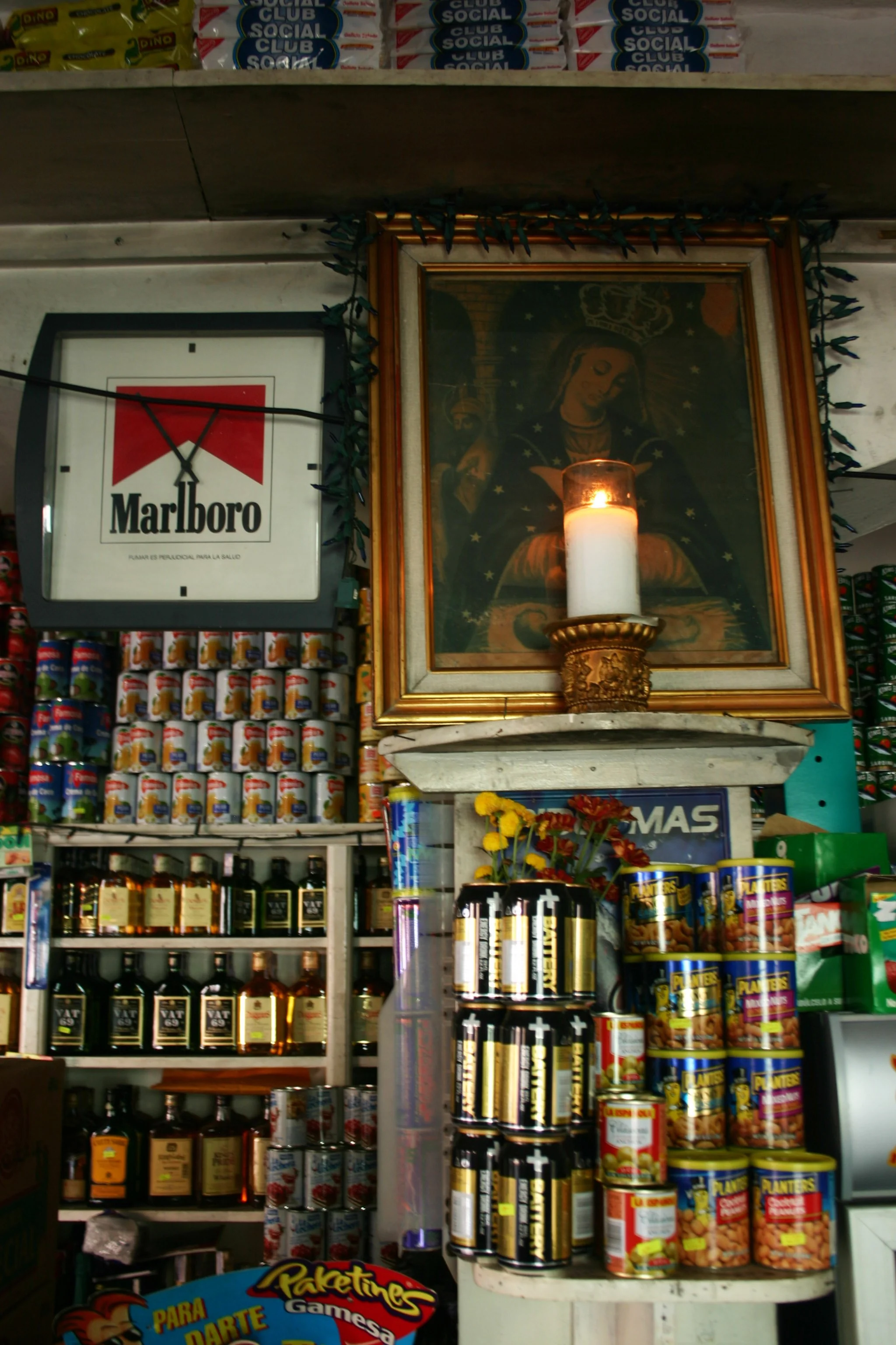 Une étagère avec différentes boîtes de conserve et bouteilles d'alcool, une image religieuse encadrée avec une bougie allumée devant, des fleurs artificielles, un panneau publicitaire pour de la marque Marlboro et un panneau dessus avec la mention 'S