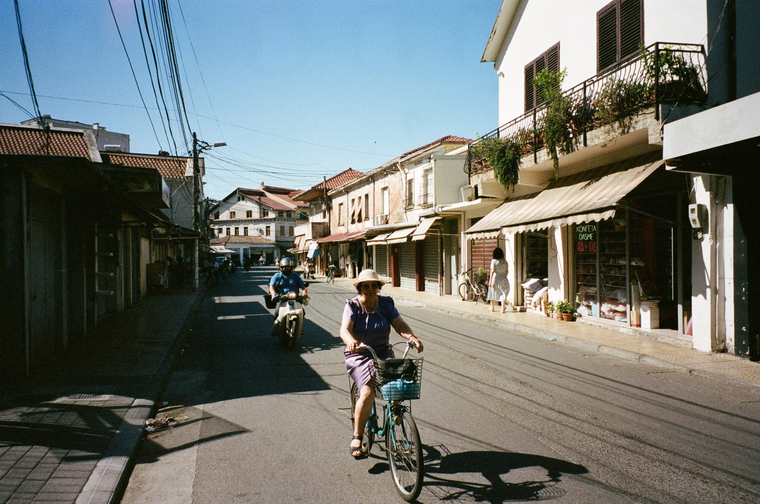 Une rue de ville avec des boutiques et des personnes, dont une femme à vélo portant un chapeau, sous un ciel bleu clair.