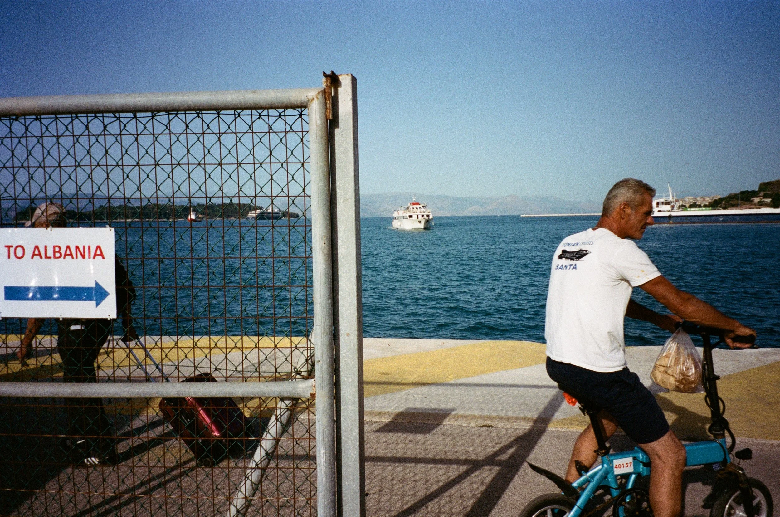 Homme âgé faisant du vélo électrique près de la mer, avec bateau à l'horizon et panneau indiquant le chemin vers l'Albanie