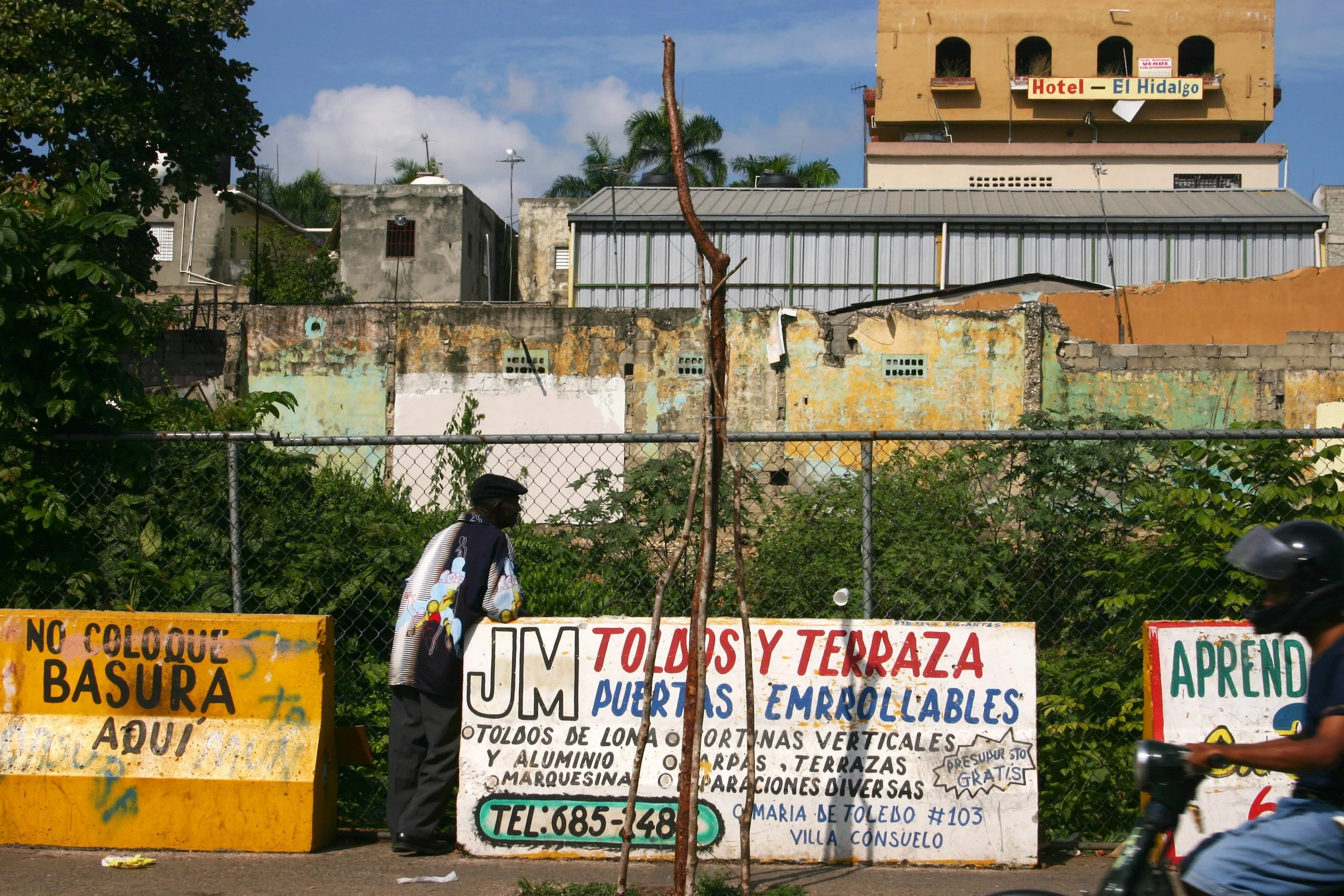 Vente de ferro-volet, portes et terrasses en métal, avec des affiches publicitaires colorées sur le trottoir et un mur en arrière-plan avec des bâtiments anciens.