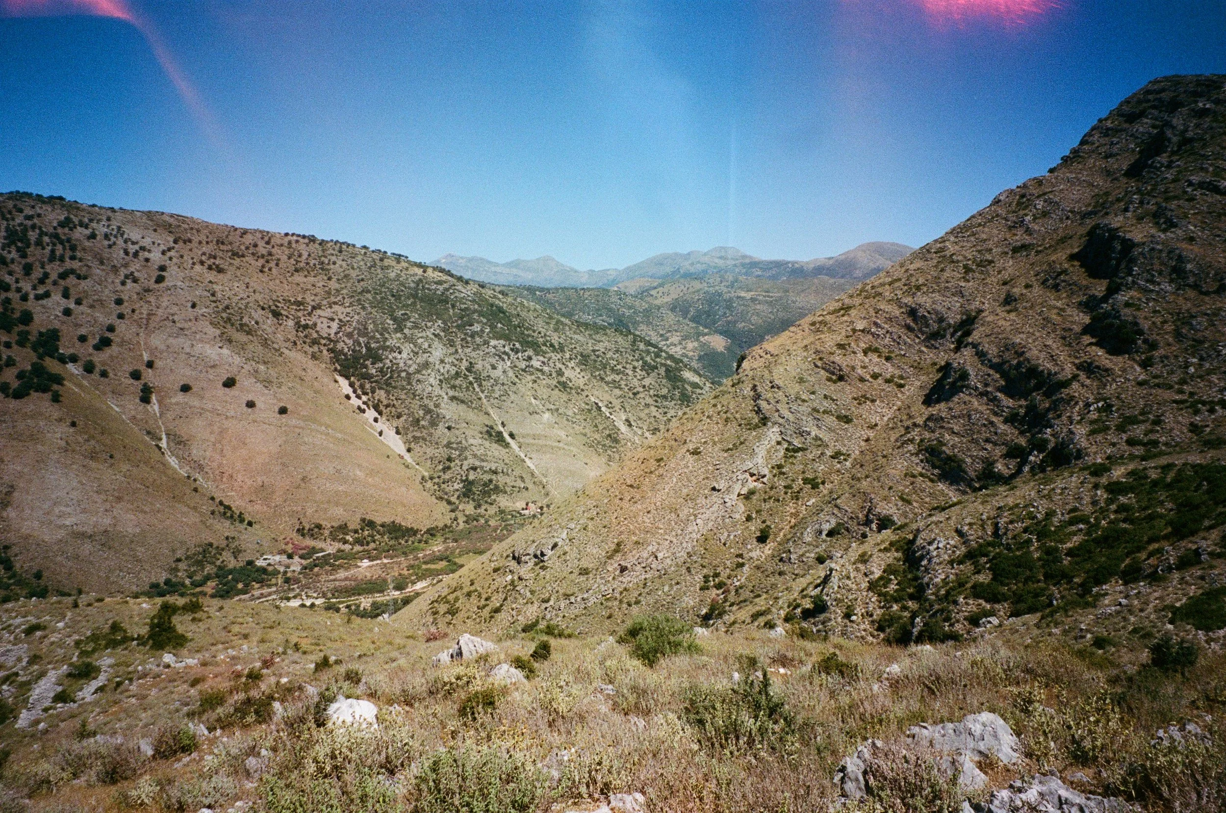 Vallée montagneuse aride avec peu de végétation et de petites maisons au fond, sous un ciel bleu clair.