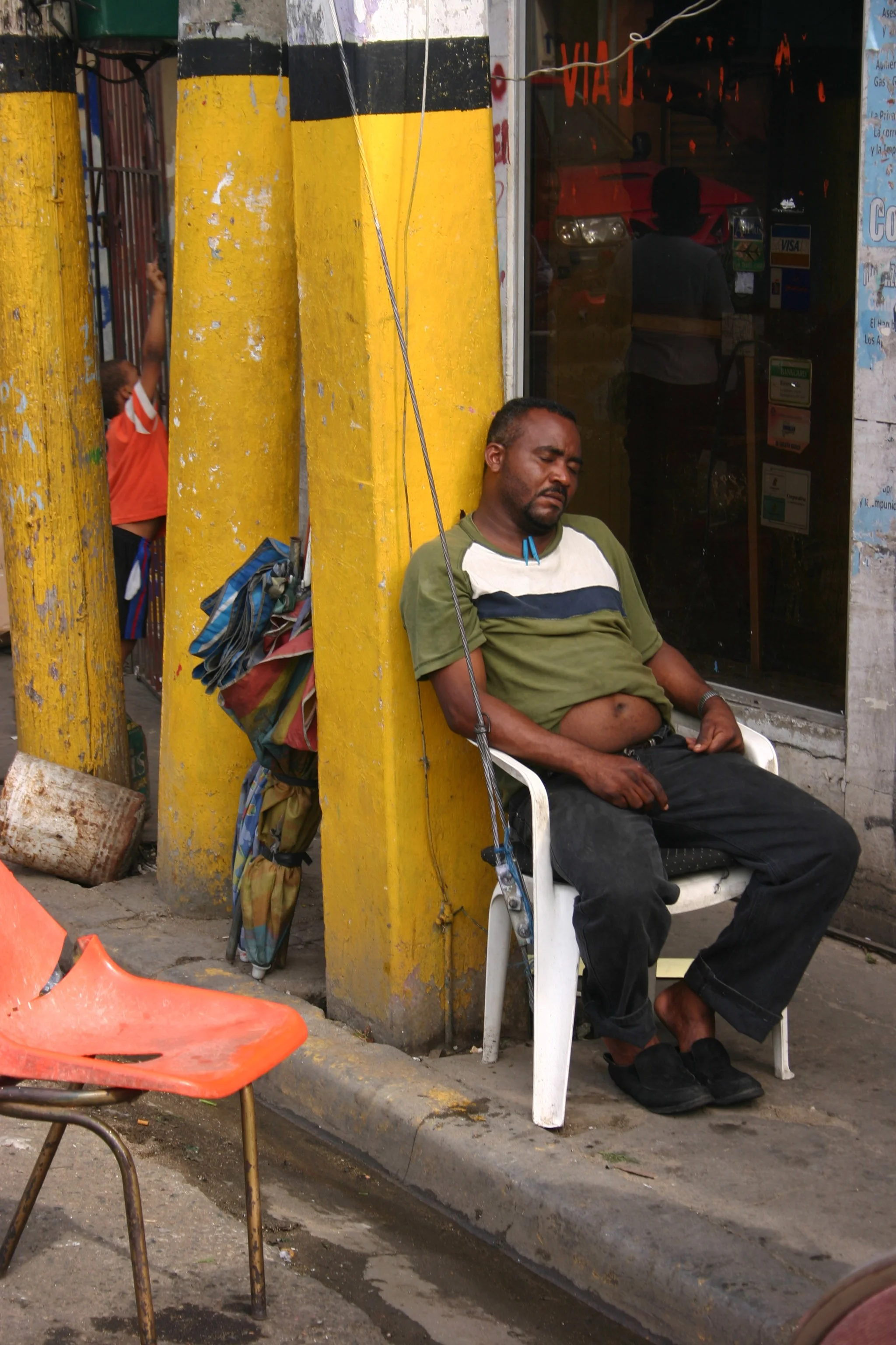 Un homme endormi assis sur une chaise en plastique blanche devant un bâtiment avec des piliers jaunes. Un garçon en orange joue derrière la barrière à gauche.
