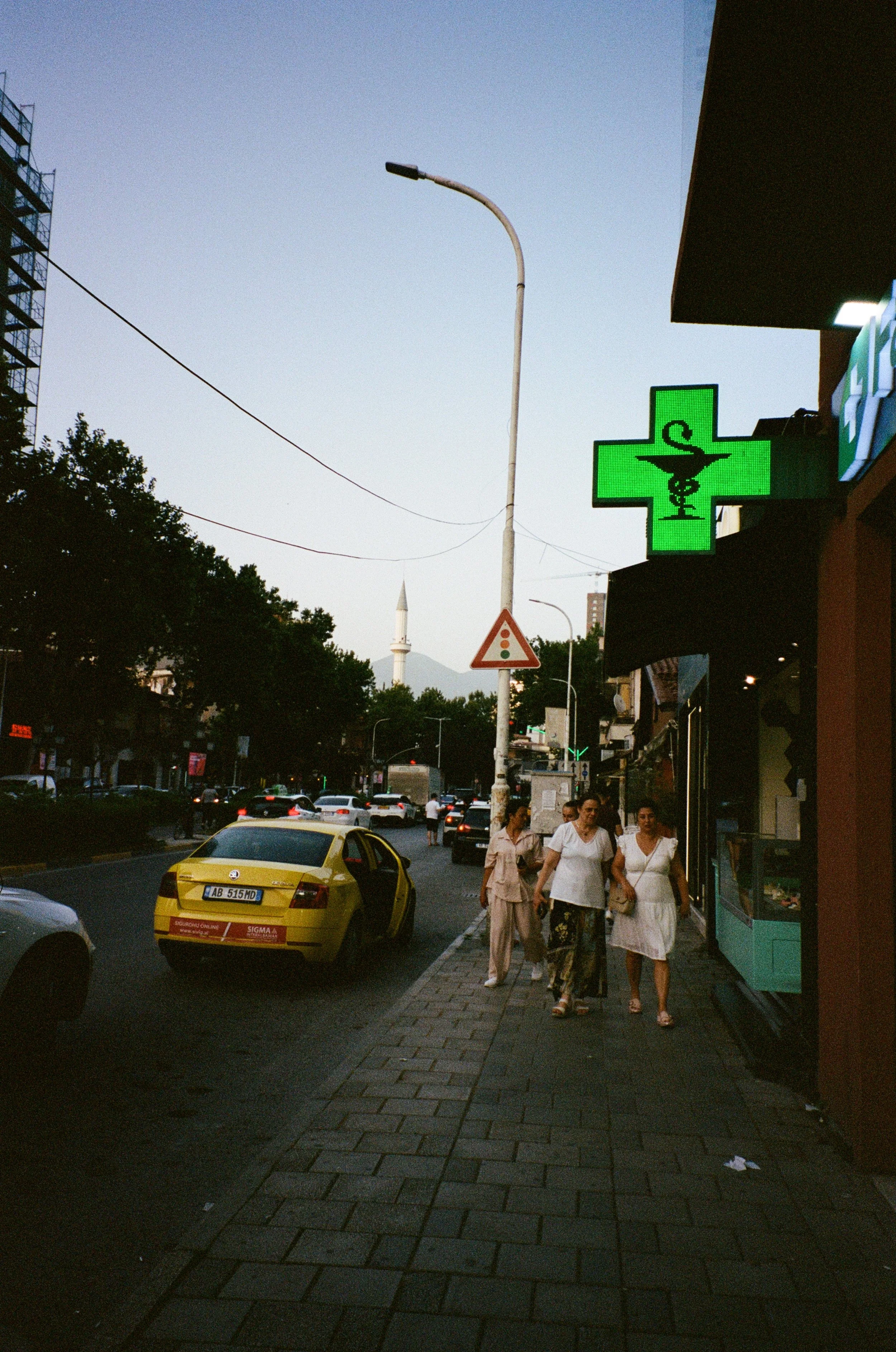 Une rue urbaine en fin d'après-midi avec des personnes marchant sur le trottoir, une voiture jaune à côté, un panneau de pharmacie lumineux vert avec une croix, un minaret en arrière-plan, et des bâtiments modernes.