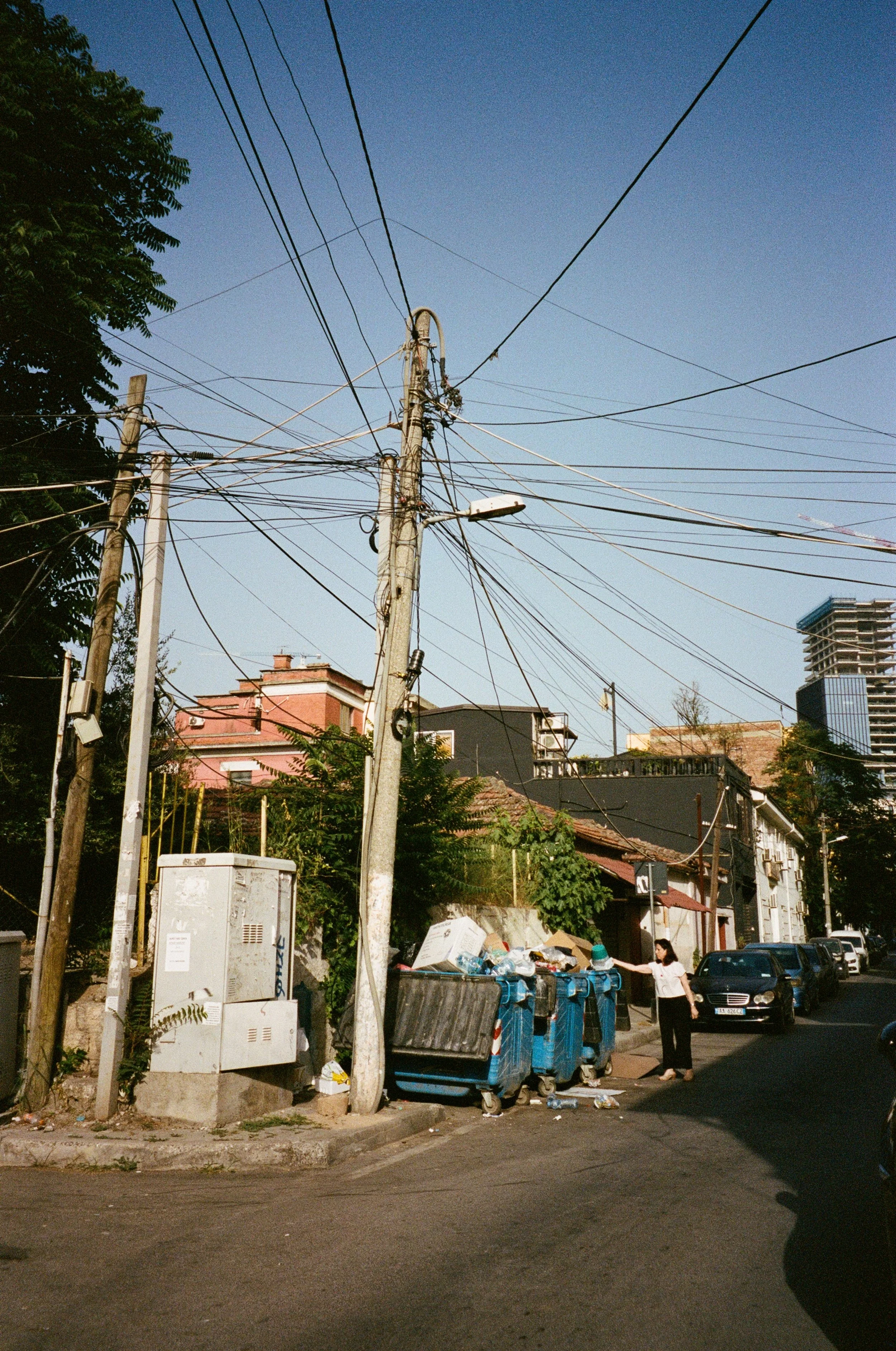 Une rue urbaine avec plusieurs poubelles, des câbles électriques suspendus, un poteau électrique, une femme qui jette quelque chose, et des bâtiments résidentiels en arrière-plan.