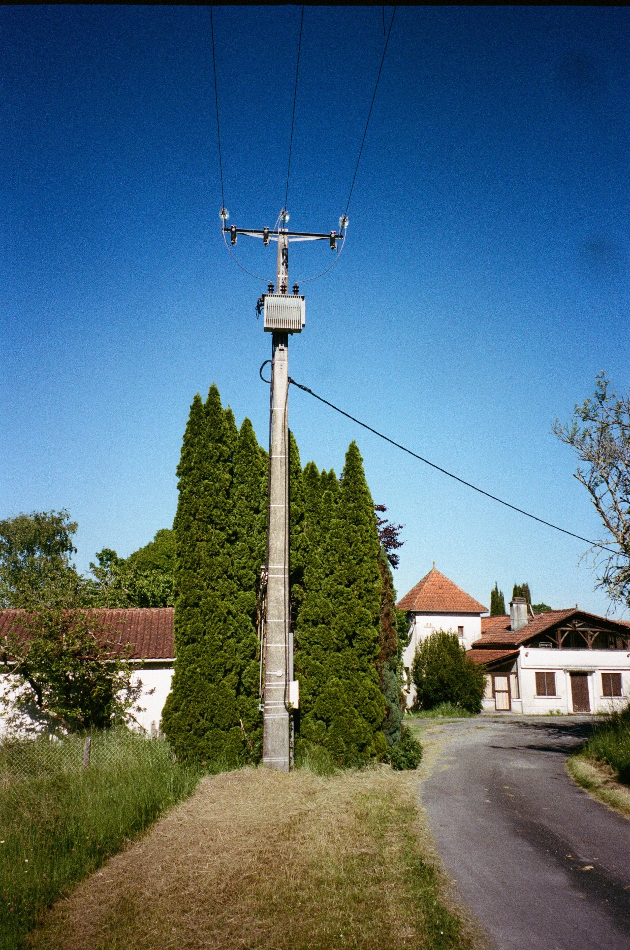 Un poteau électrique en béton avec des lignes électriques sous un ciel bleu clair, entouré d'arbres verts et de maisons résidentielles dans un village.