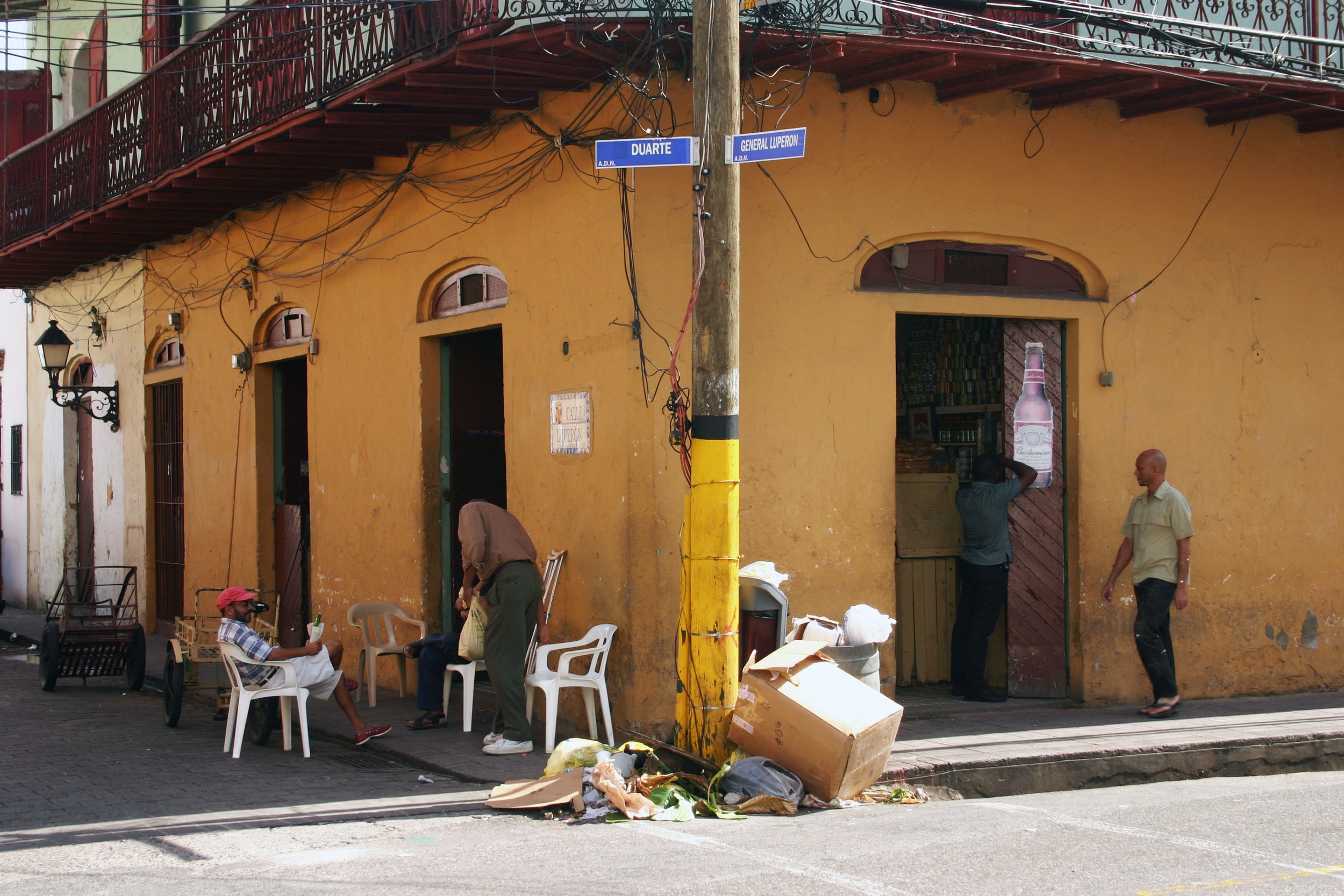 Rue avec des personnes assises et discutant, devant un bâtiment jaune avec des portes ouvertes, autour de poubelles et de déchets. Deux hommes près de la porte, un homme avec une casquette rouge assis, d'autres personnes discutant ou regardant.