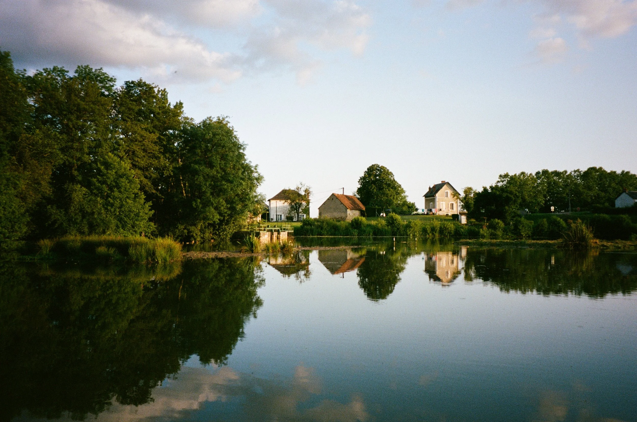 Maisons près d'un lac paisible avec des arbres, reflet dans l'eau, ciel clair avec quelques nuages, en fin de journée ou au début de soirée.
