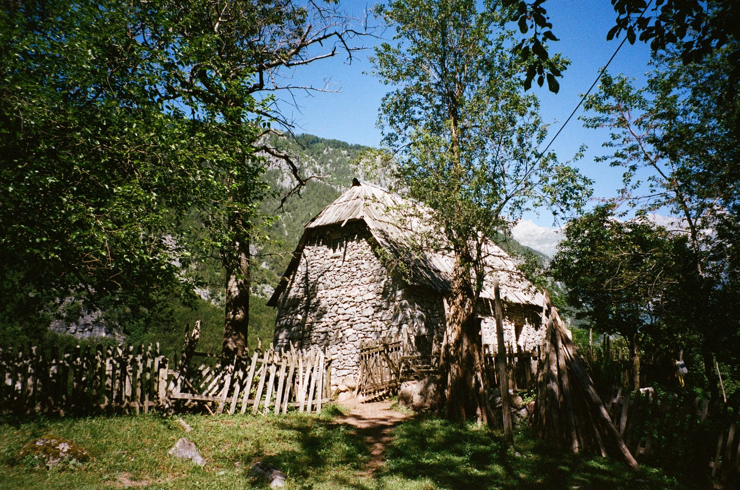 Petite maison en pierre avec toit en bois, entourée d'arbres et de grillages en bois, sous un ciel bleu, dans un environnement rural montagneux.