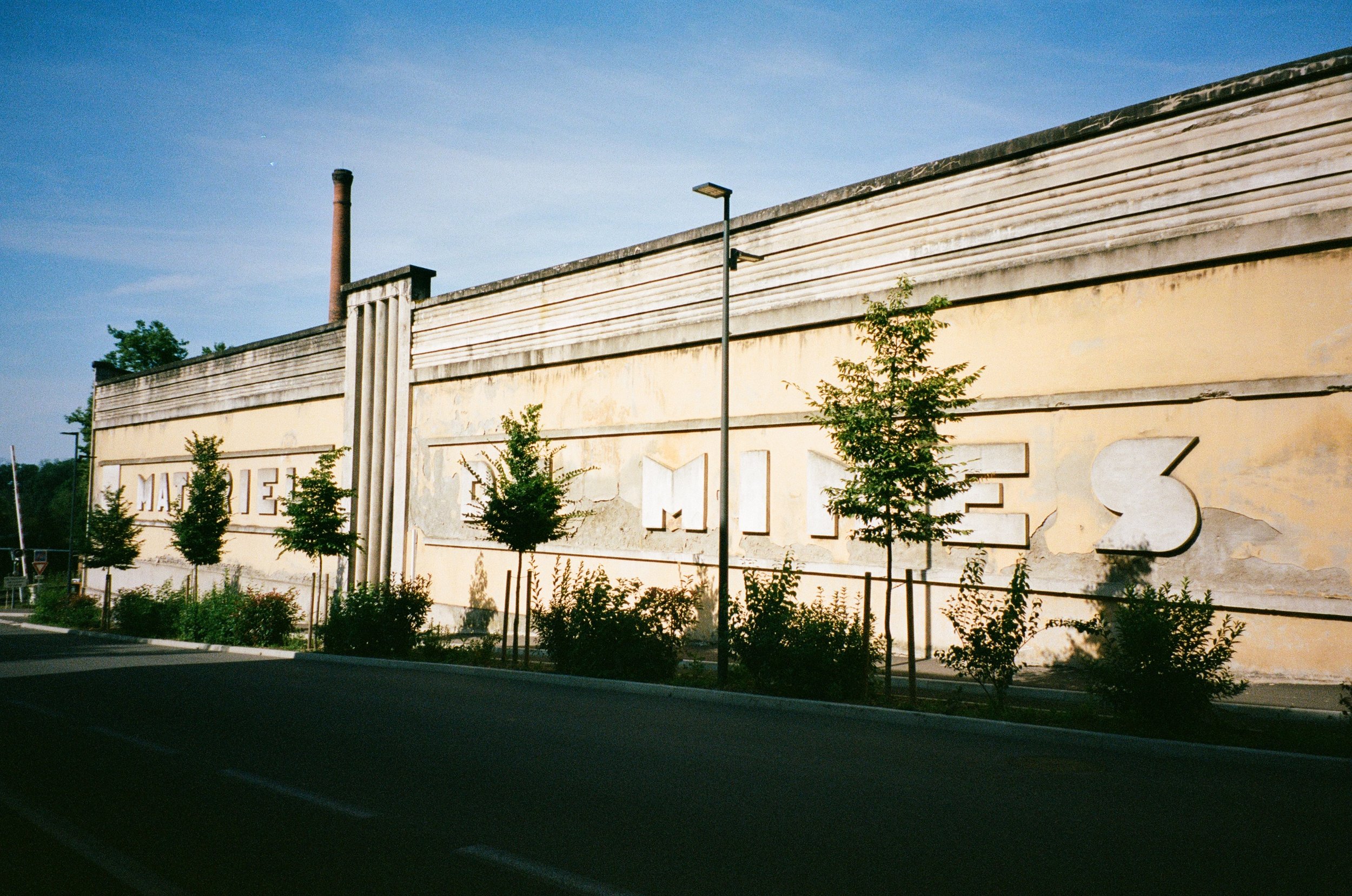 Mur avec inscription en relief partiellement visible, entouré d'arbres et de jeunes arbustes, avec un lampadaire et une cheminée en arrière-plan, sous un ciel bleu clair.
