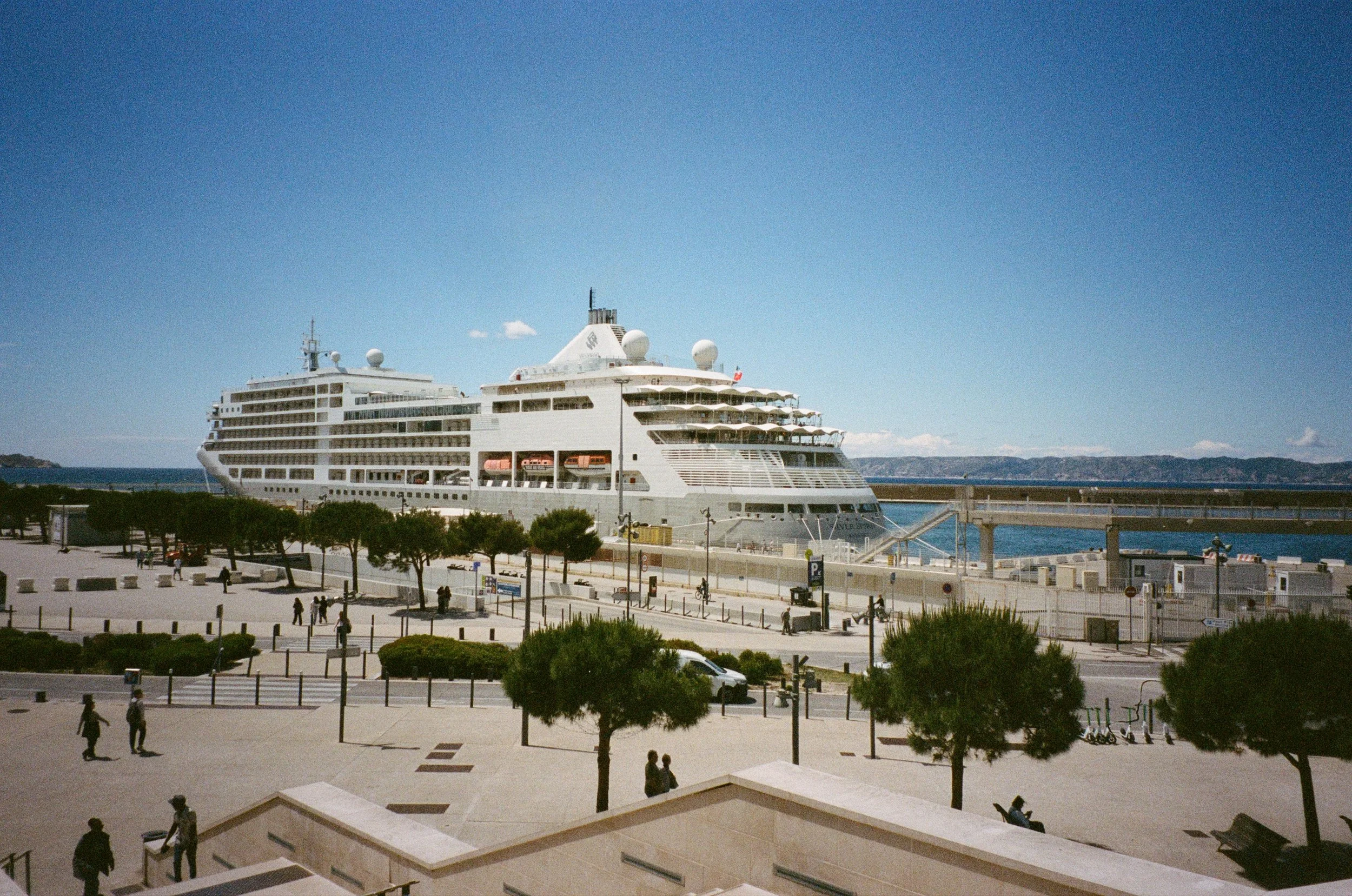 Un grand bateau de croisière blanc amarré dans un port, avec un ciel bleu et quelques nuages, entouré de quelques arbres et personnes qui se promènent.