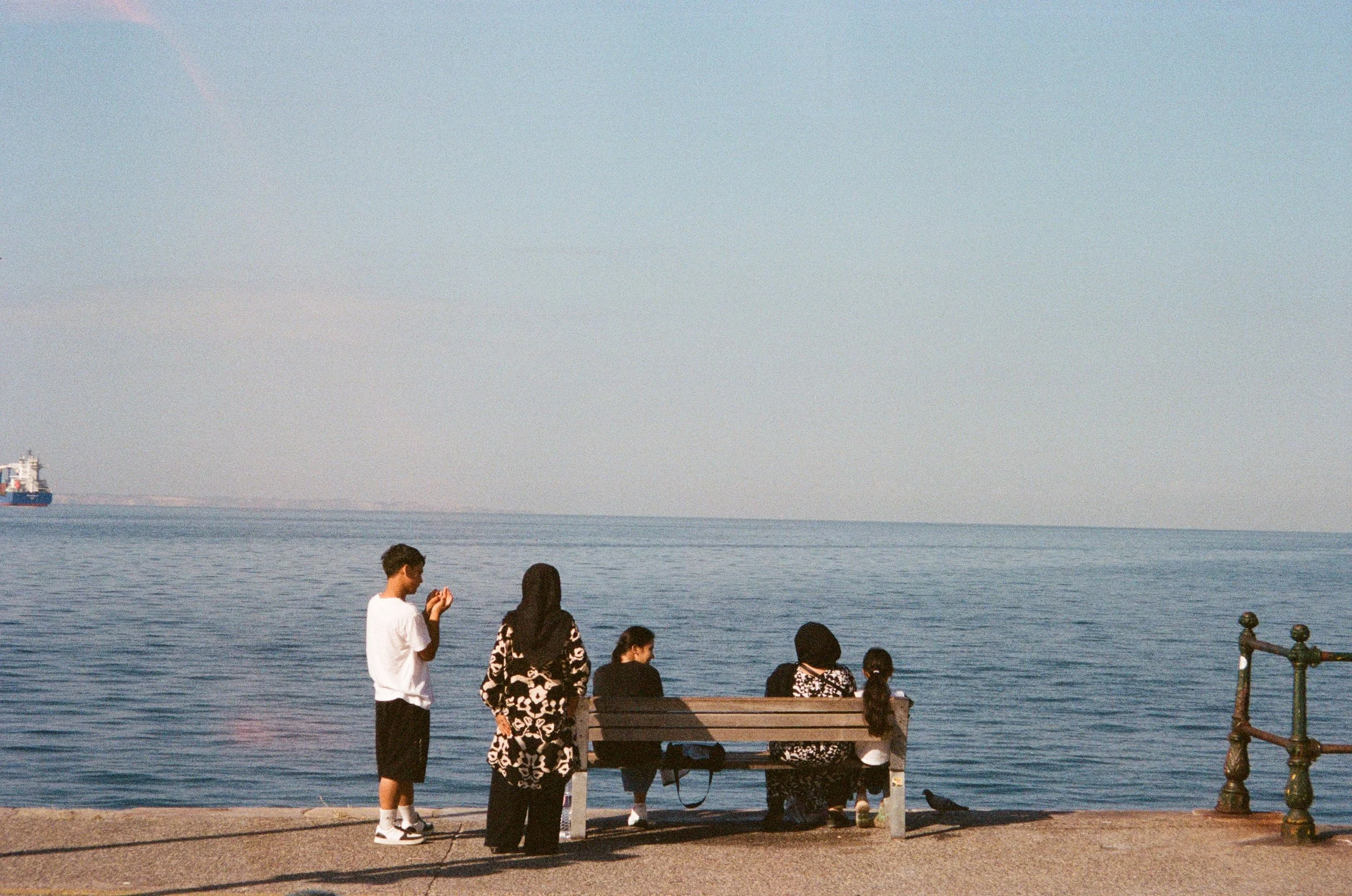 Quatre personnes près d'une mer, assises sur un banc en train de regarder l'eau, une personne debout à côté prenant une photo ou regardant leur téléphone, un bateau en arrière-plan, ciel clair.