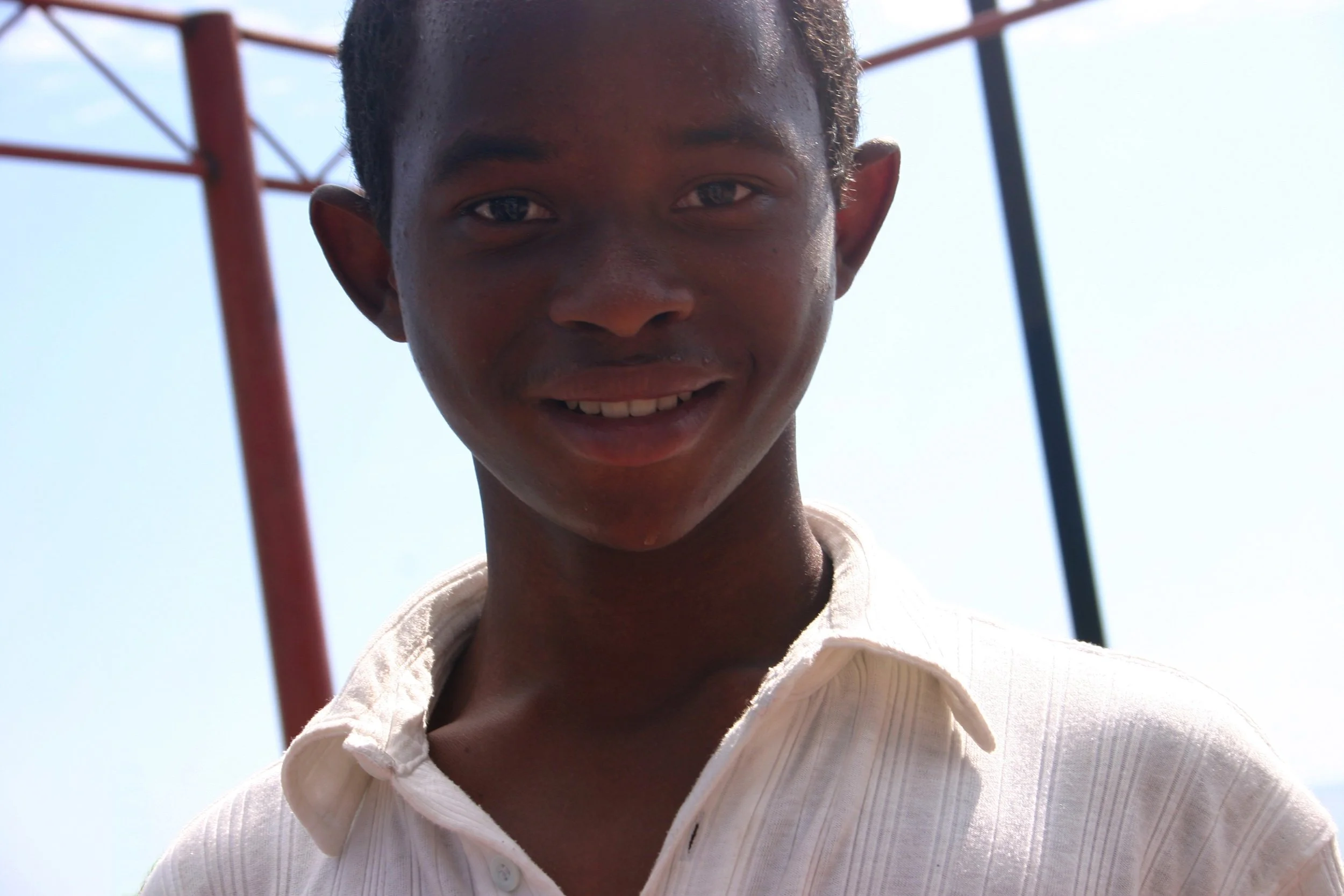Un garçon souriant portant une chemise beige, en extérieur sous un ciel clair.