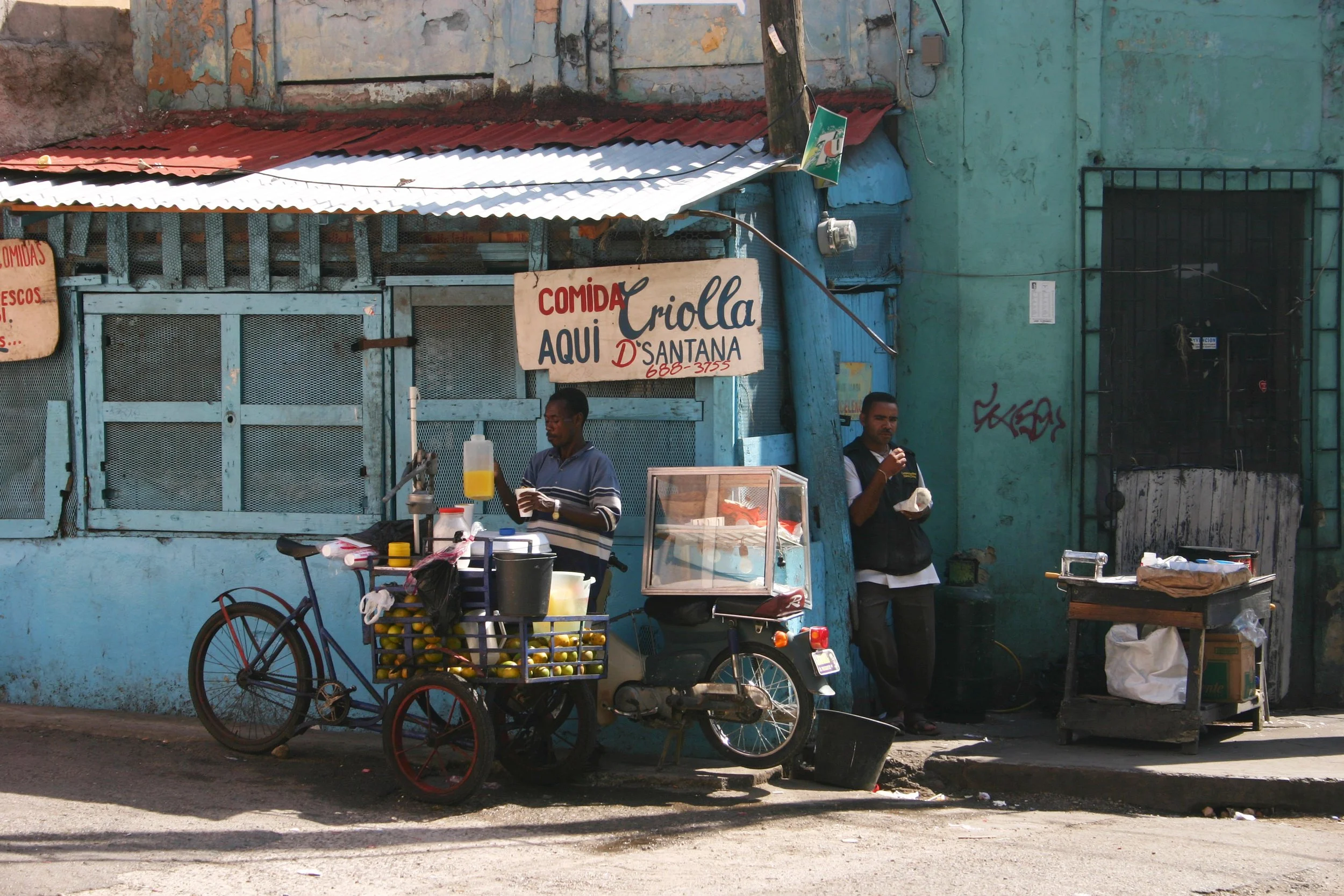 Deux hommes vendant des citrons dans une rue en face d'un petit stand de nourriture avec un signe indiquant 'Comida Criolla Aquí D'Santana'.