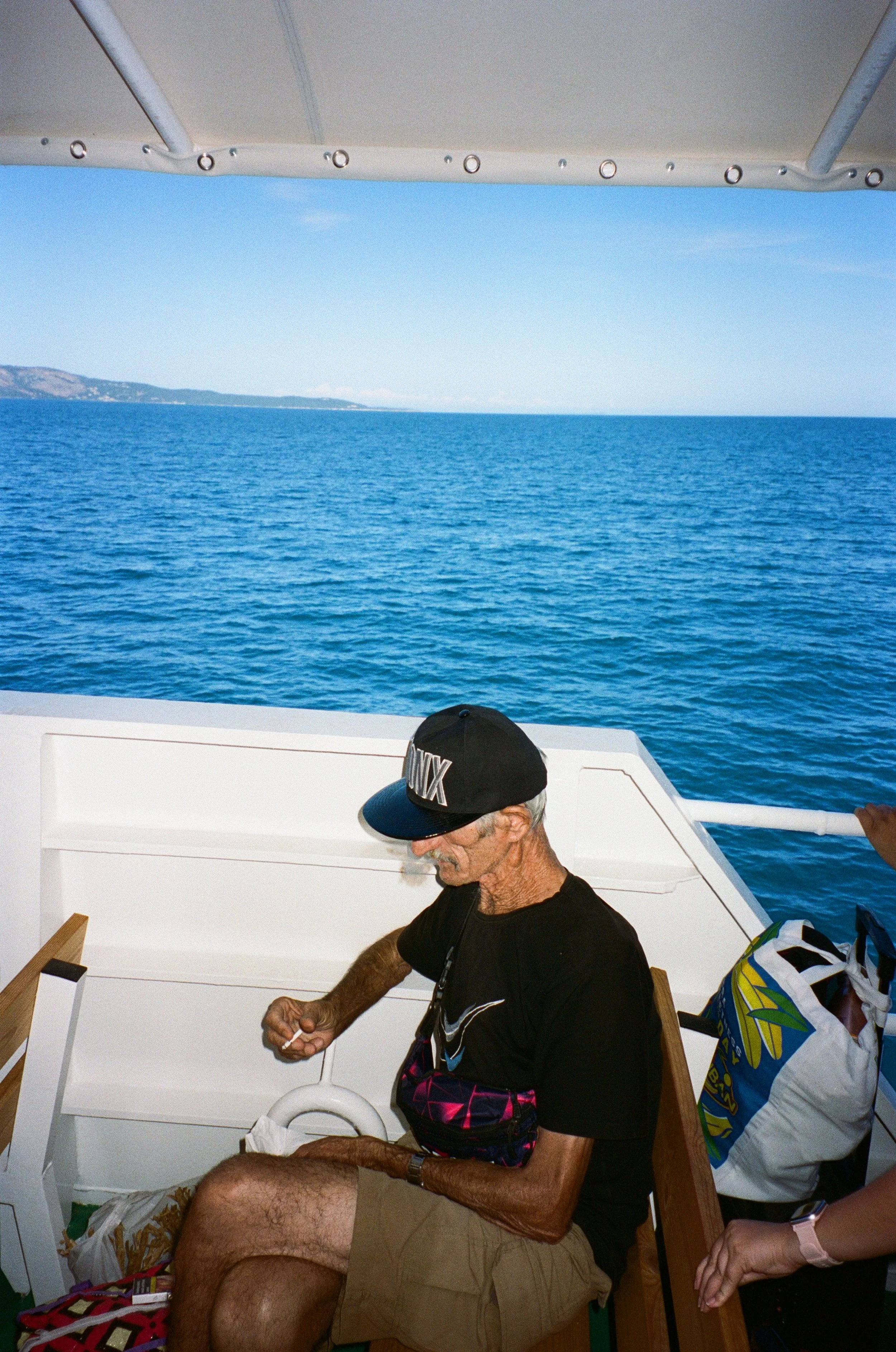 Un homme âgé avec une casquette noire, un t-shirt noir, et un short beige, assis sur un bateau en mer, avec un paysage côtier à l'horizon.