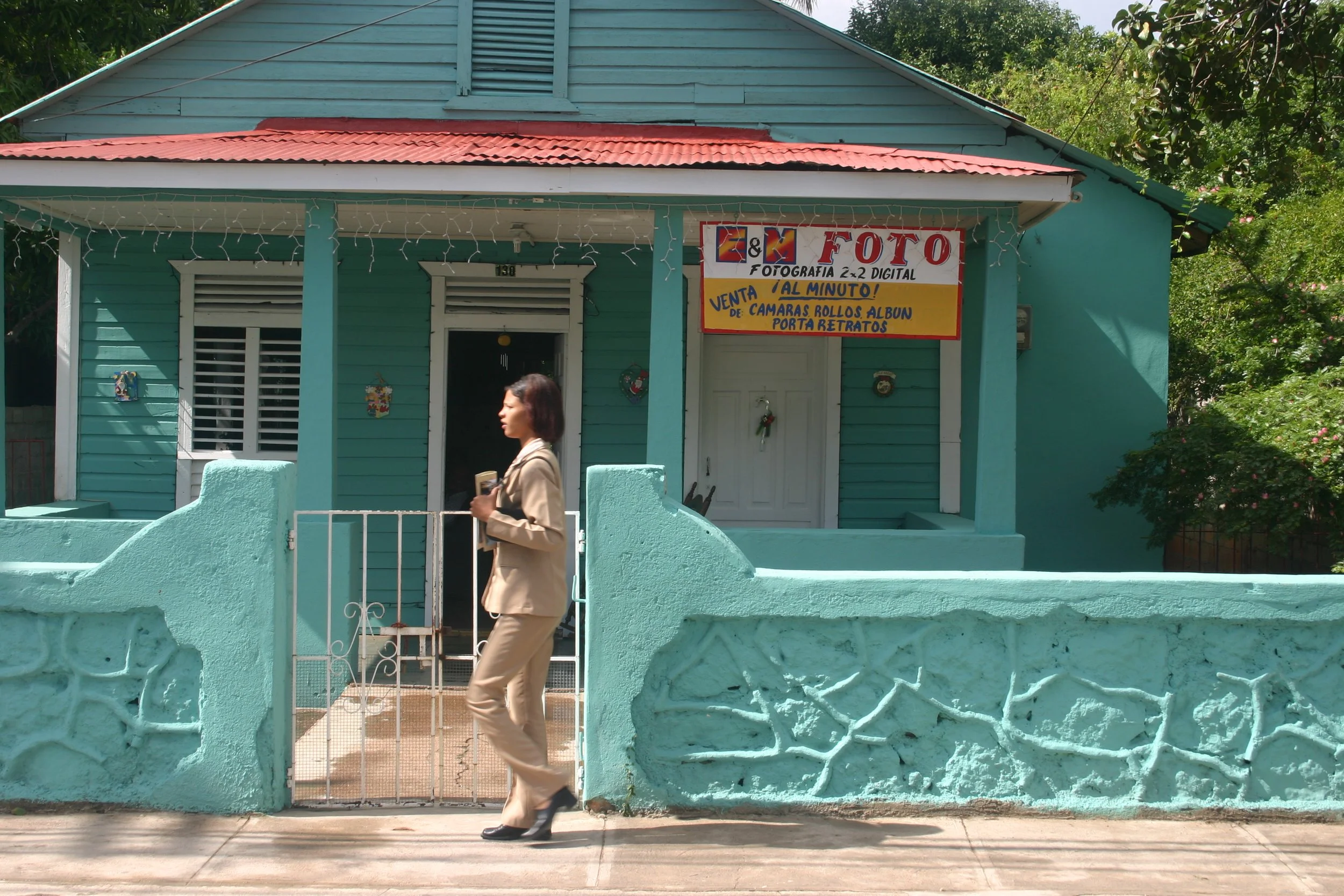 Une femme marchant devant une maison en bois peinte en turquoise, avec un panneau publicitaire pour une boutique de photographie, décoration florale et un petit mur en pierre.