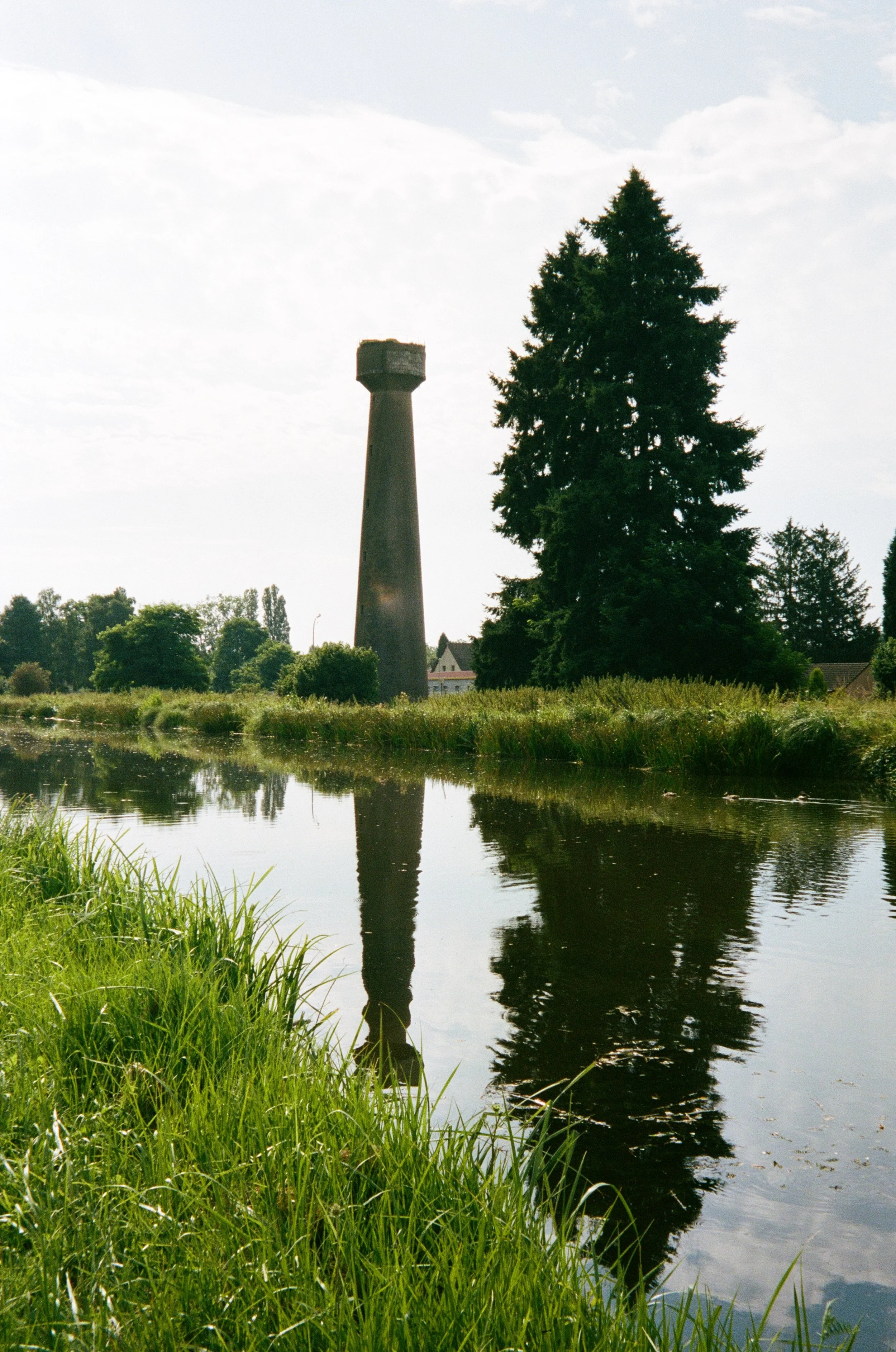 Une tour en béton à côté d'un grand arbre près d'un étang, avec des reflets dans l'eau, sous un ciel partiellement nuageux.