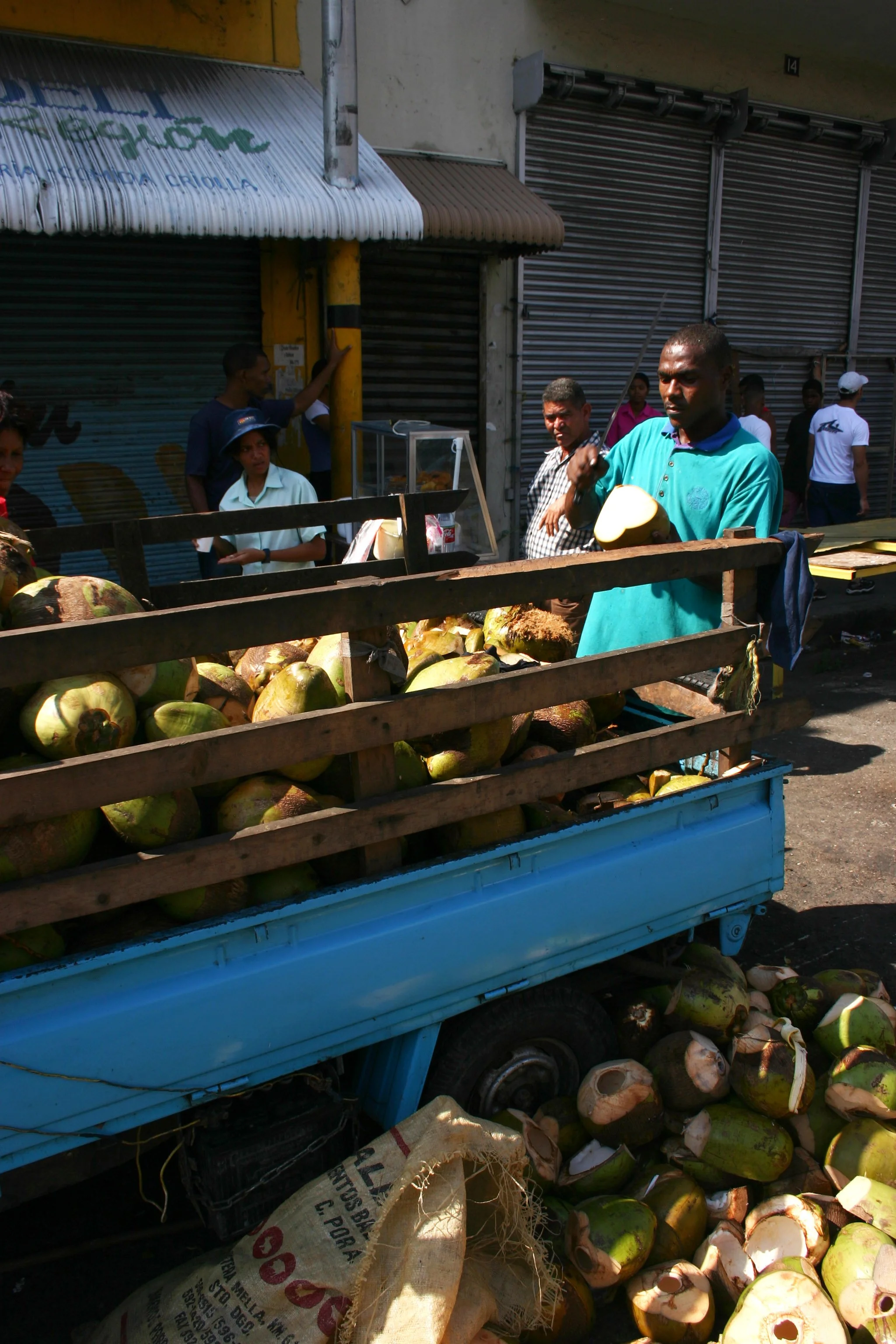 Marché en plein air avec un vendeur vendant des noix de coco dans une remorque en métal bleue, plusieurs personnes en arrière-plan, devant des boutiques avec des volets métalliques.