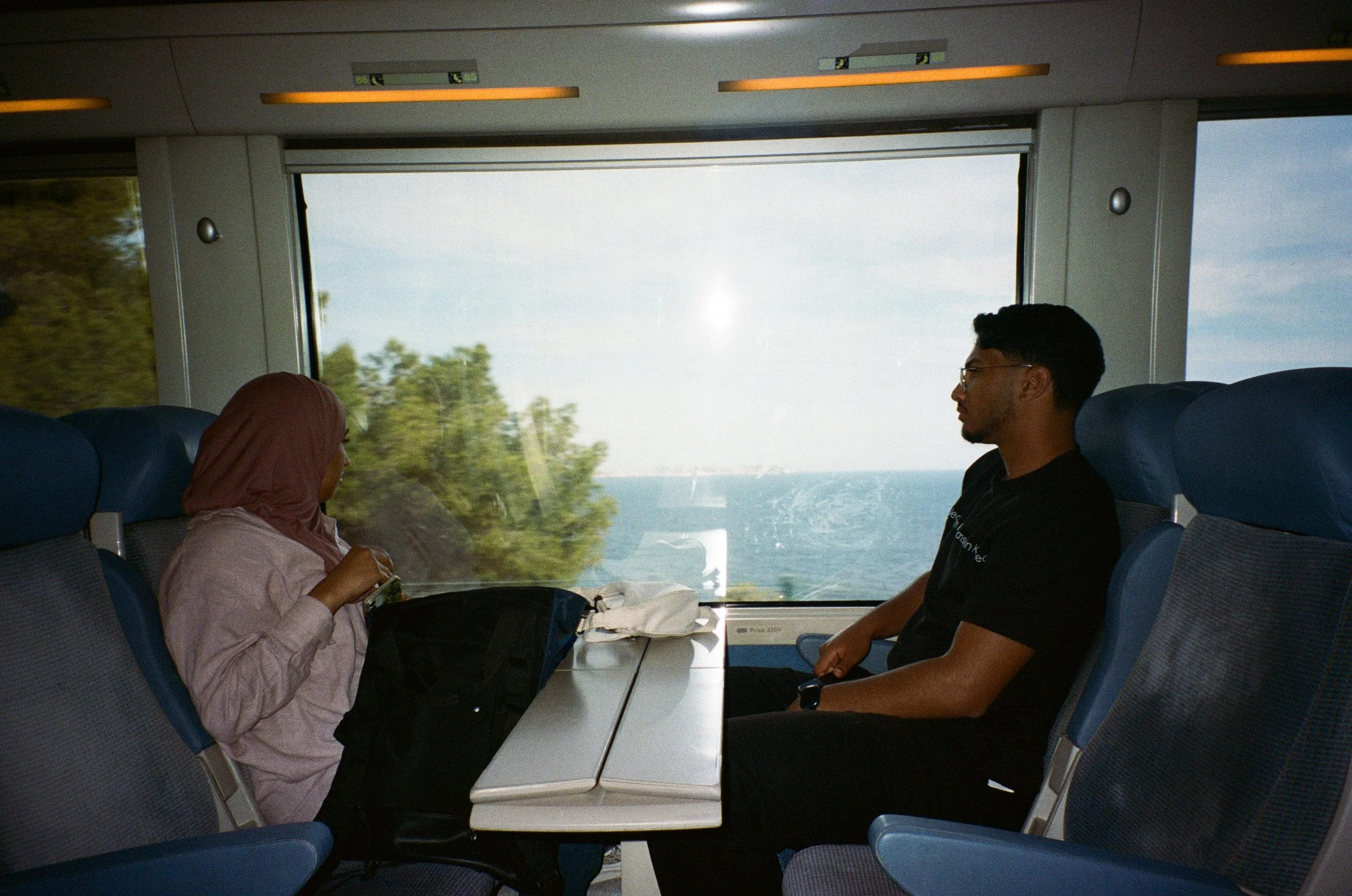Deux personnes assises face à face dans un train, parlant, avec vue sur la mer et la nature à travers la fenêtre.