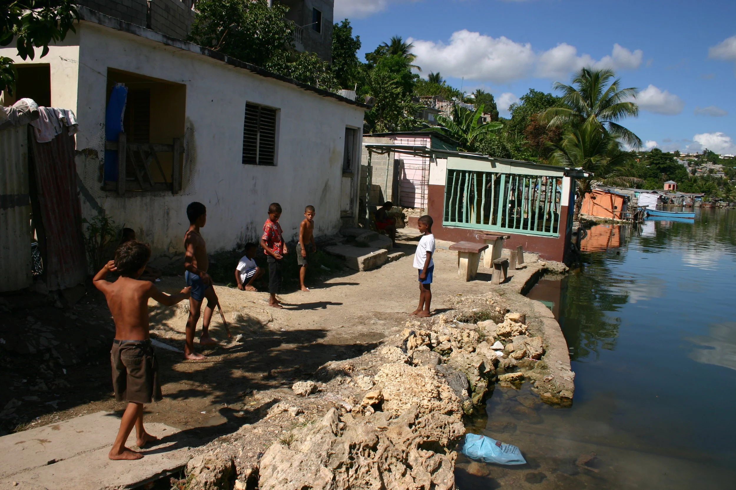 Enfants jouent près d'une rivière devant des maisons modestes dans un quartier en hauteur, avec des palmiers en arrière-plan, sous un ciel en partie nuageux.