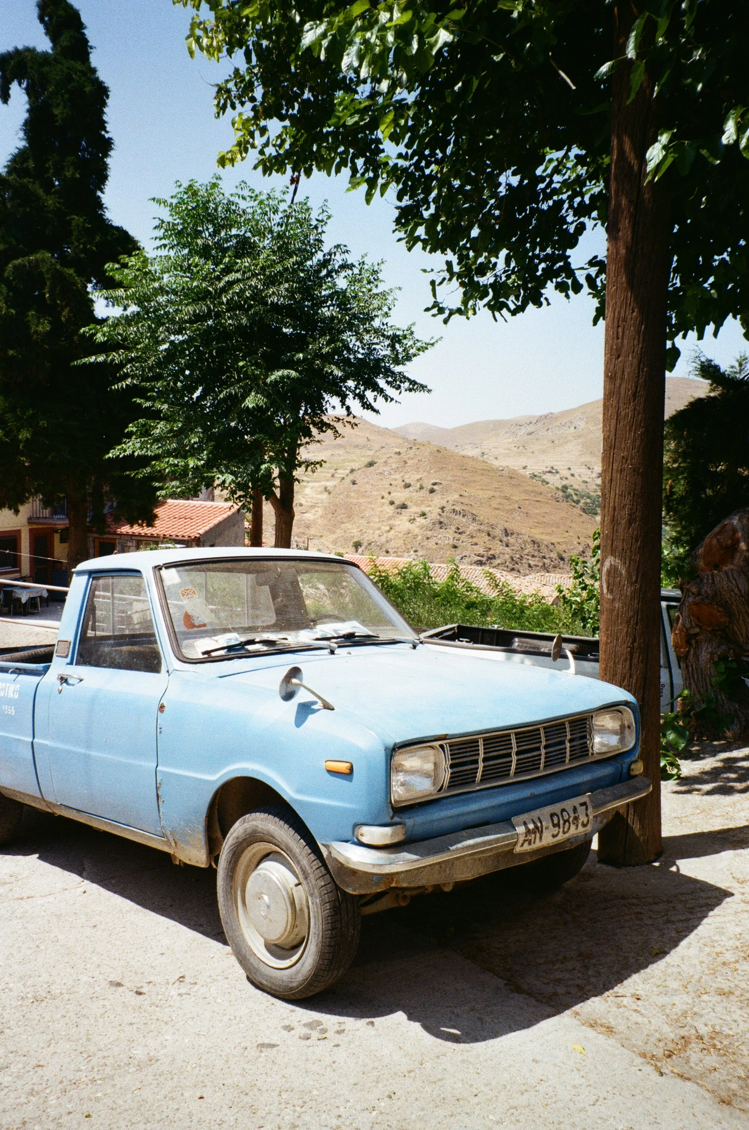 Voiture ancienne bleue stationnée sous un arbre avec des montagnes en arrière-plan.