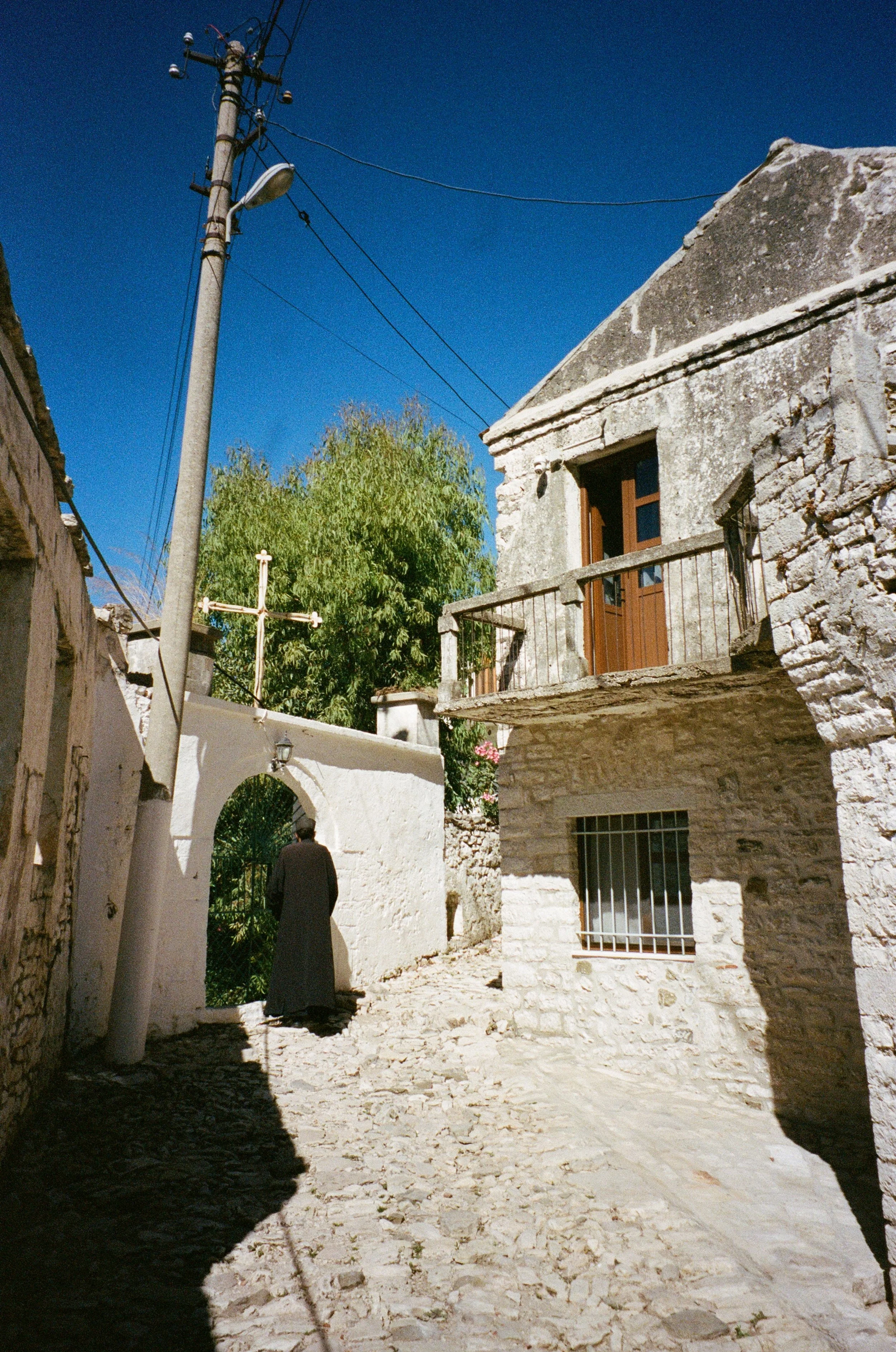 Une rue pavée dans un village avec des pierres, une personne en noir, une maison en pierre avec un balcon, un arbre et un ciel bleu.