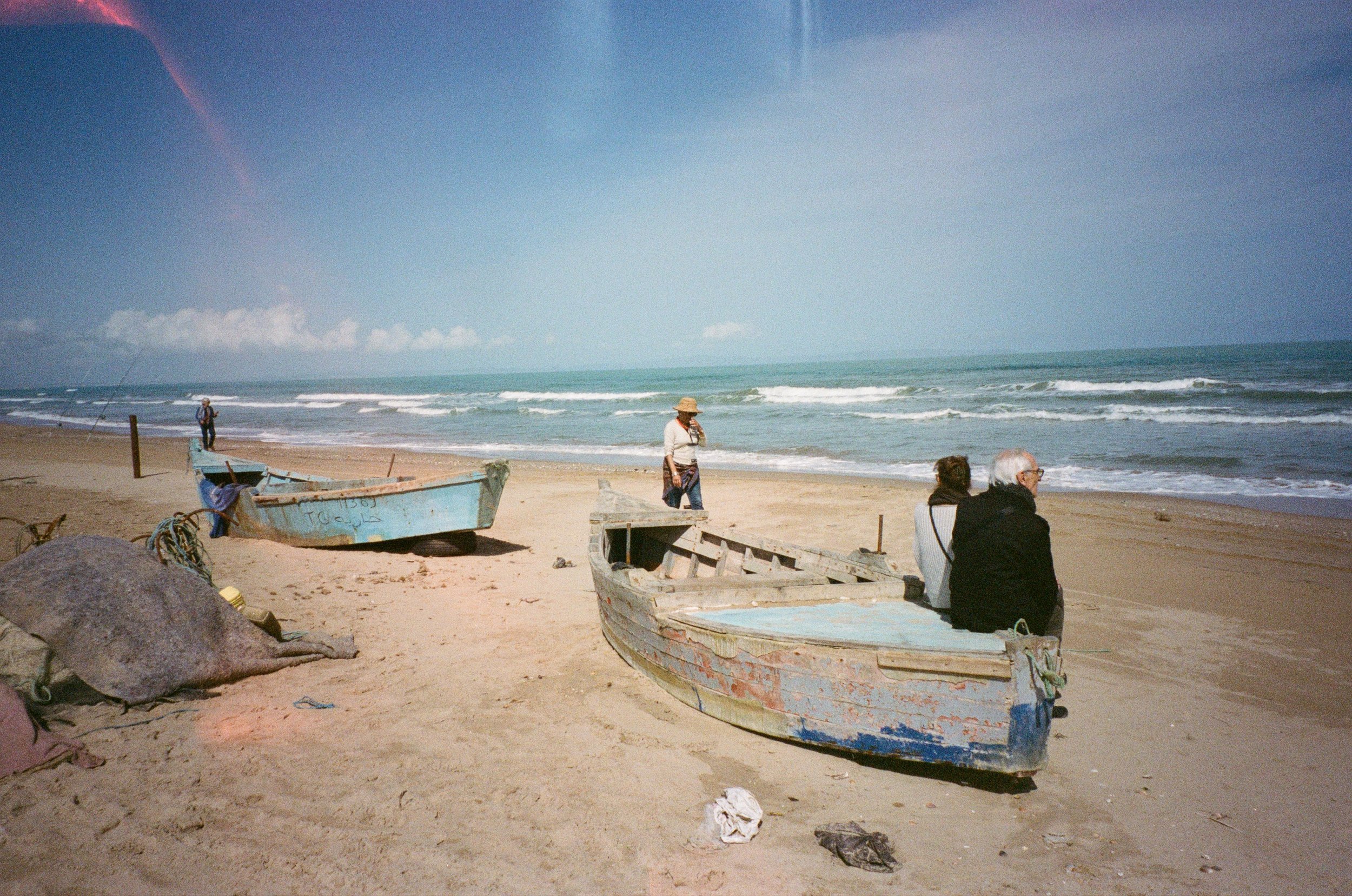 Plage avec deux bateaux en bois abandonnés, trois personnes, deux assises sur un bateau et une marchant, la mer en arrière-plan sous un ciel bleu avec quelques nuages.