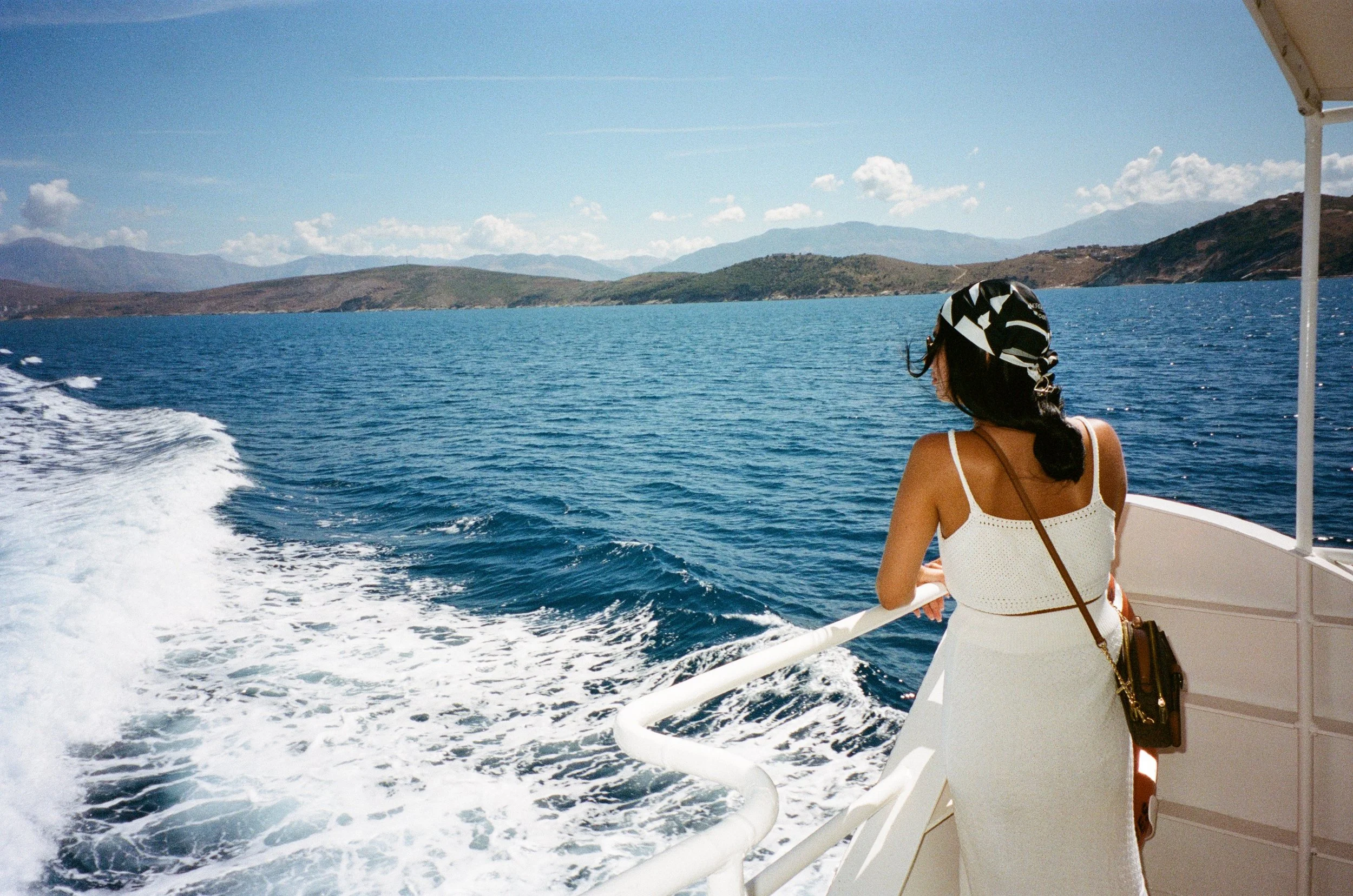 Femme regardant la mer depuis un bateau ensoleillé, avec des collines et des montagnes en arrière-plan.