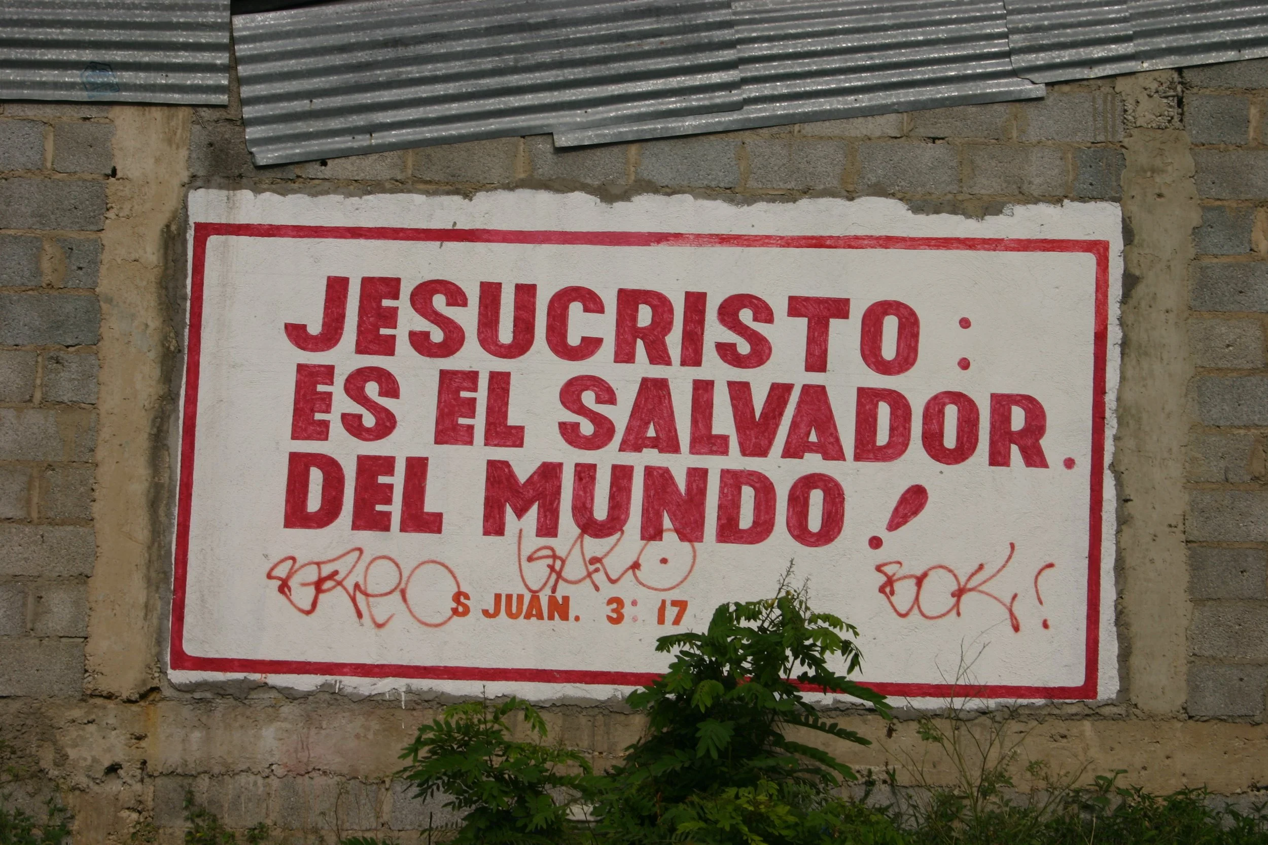 Une affiche peinte sur un mur en briques, avec une citerne de tôle au sommet. L'affiche demande si Jésus-Christ est le Sauveur du monde.