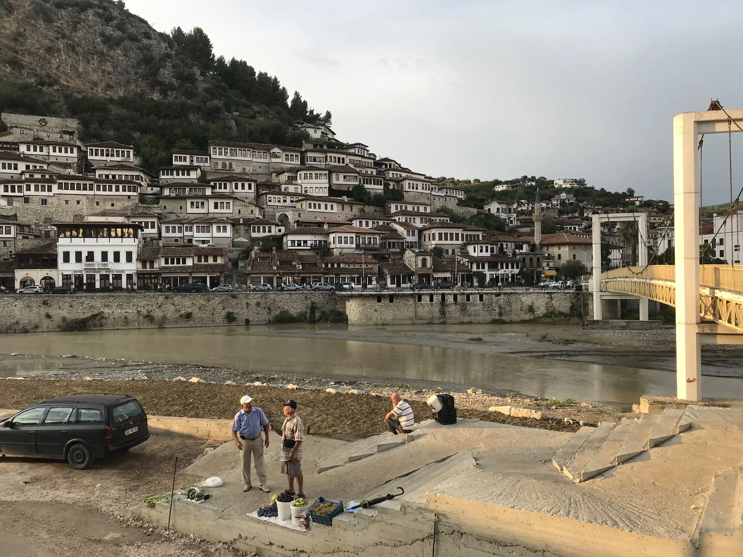 Deux hommes discutent sur un trottoir en face d'un marché de fruits. En arrière-plan, il y a une rivière, une ville avec des maisons traditionnelles en terrasses sur la colline, et un pont jaune moderne.