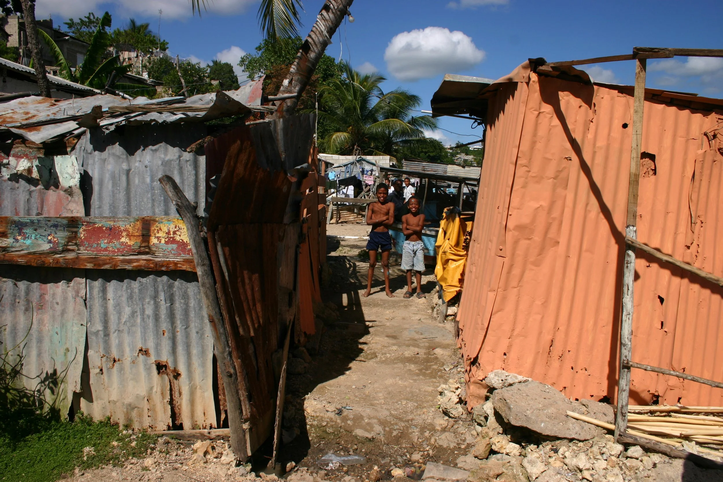 Enfants souriants dans un village avec des maisons en matériaux recyclés et en tôle ondulée, sous un ciel bleu avec quelques nuages, entouré de végétation tropicale.