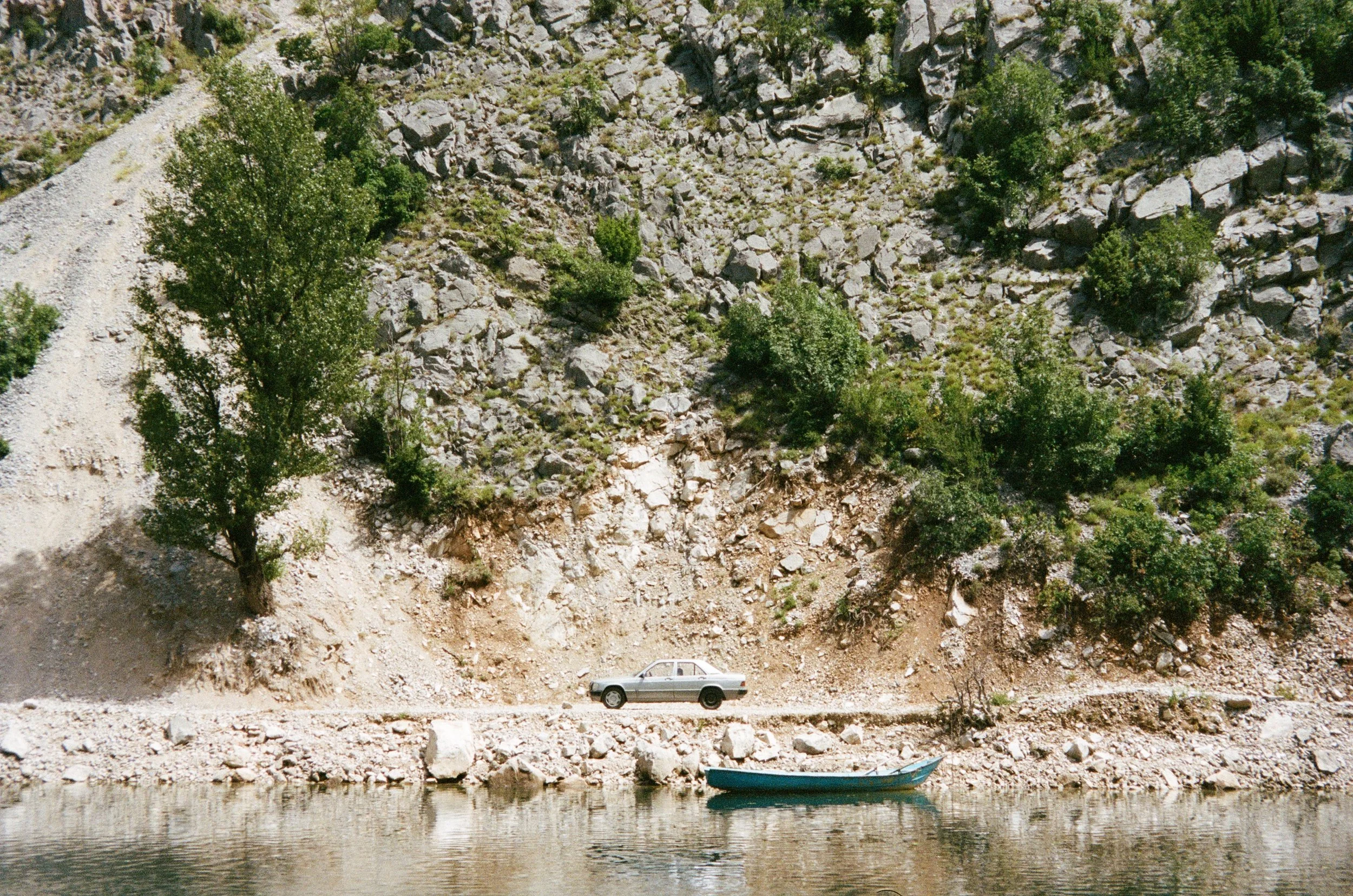 Voiture argentée stationnée sur un chemin le long d'une rivière, avec un bateau à côté, au pied d'une roche montagneuse avec des arbres verts.