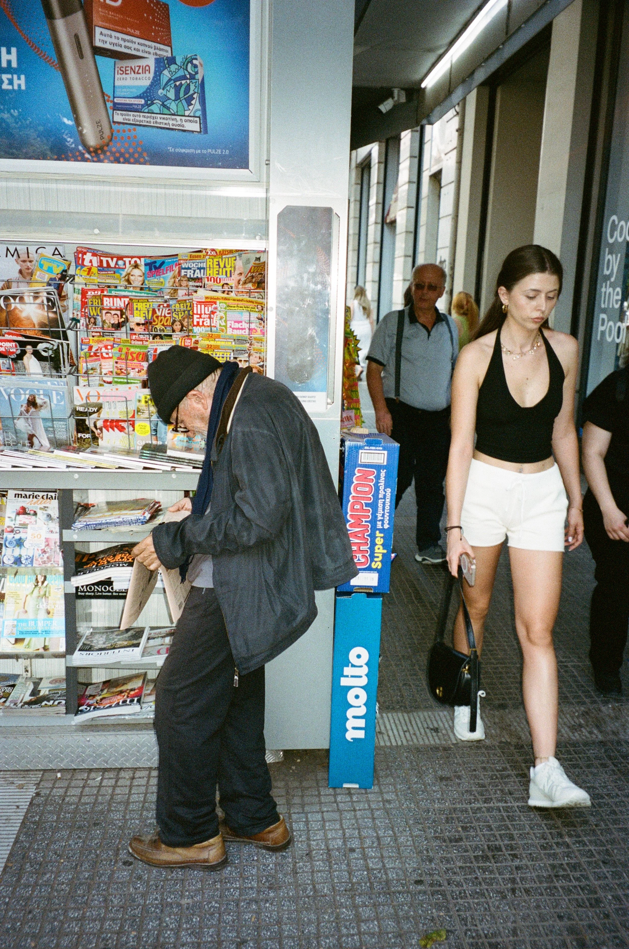 Un homme âgé portent un manteau noir et un bonnet, regardant un magazine à une cabine de magazines. Une jeune femme portant un haut noir, un short blanc et des baskets blanches marche à côté, tenant un téléphone portable et un sac noir. Dans le fond,