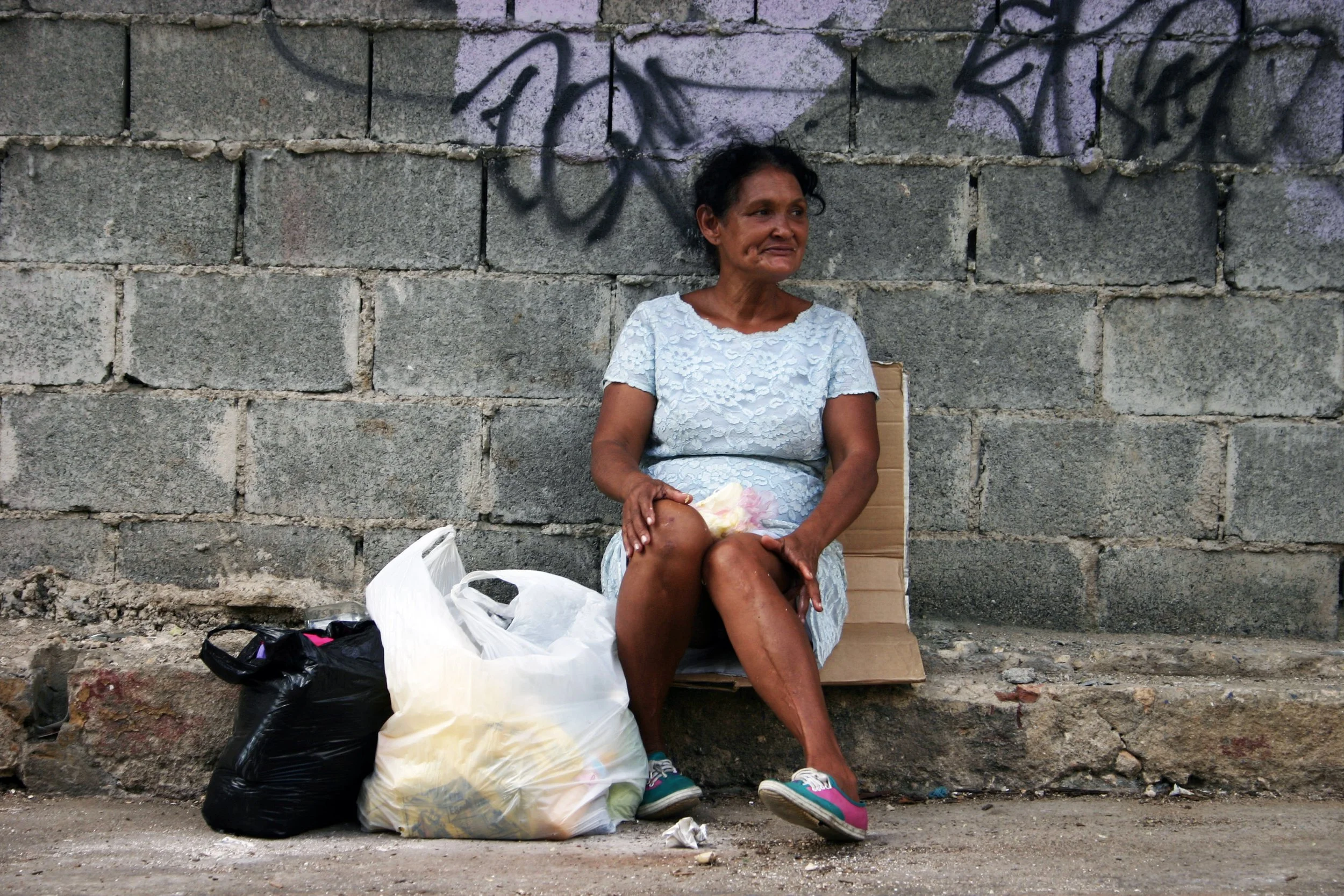 Une femme assise contre un mur en brique grise, entourée de sacs d'ordures, portant une robe bleue et des chaussures de sport roses.