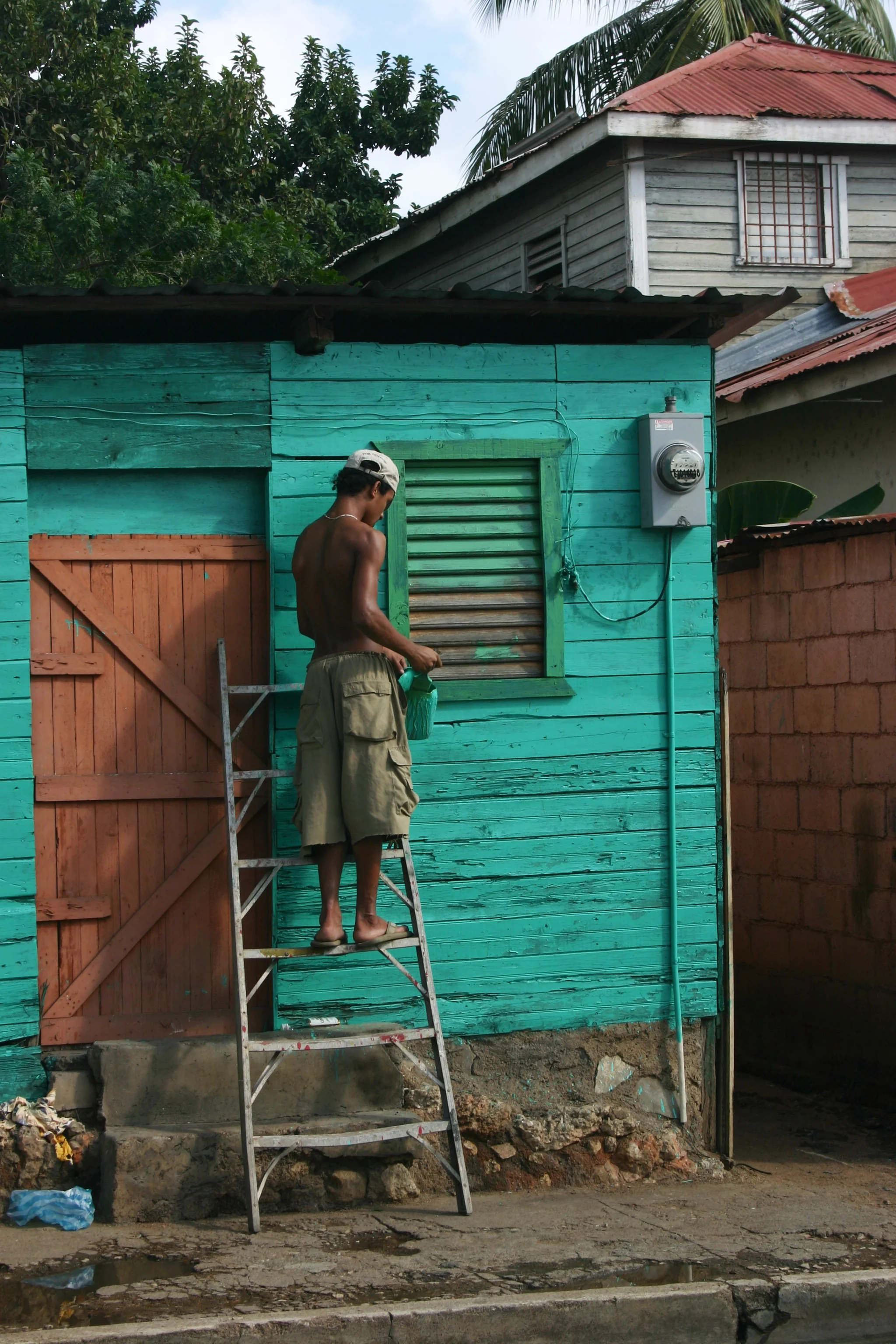 Un homme nu debout sur une échelle devant une maison en bois turquoise, tenant une bouteille verte. La maison a une fenêtre à volets et un compteur électrique à l'extérieur.