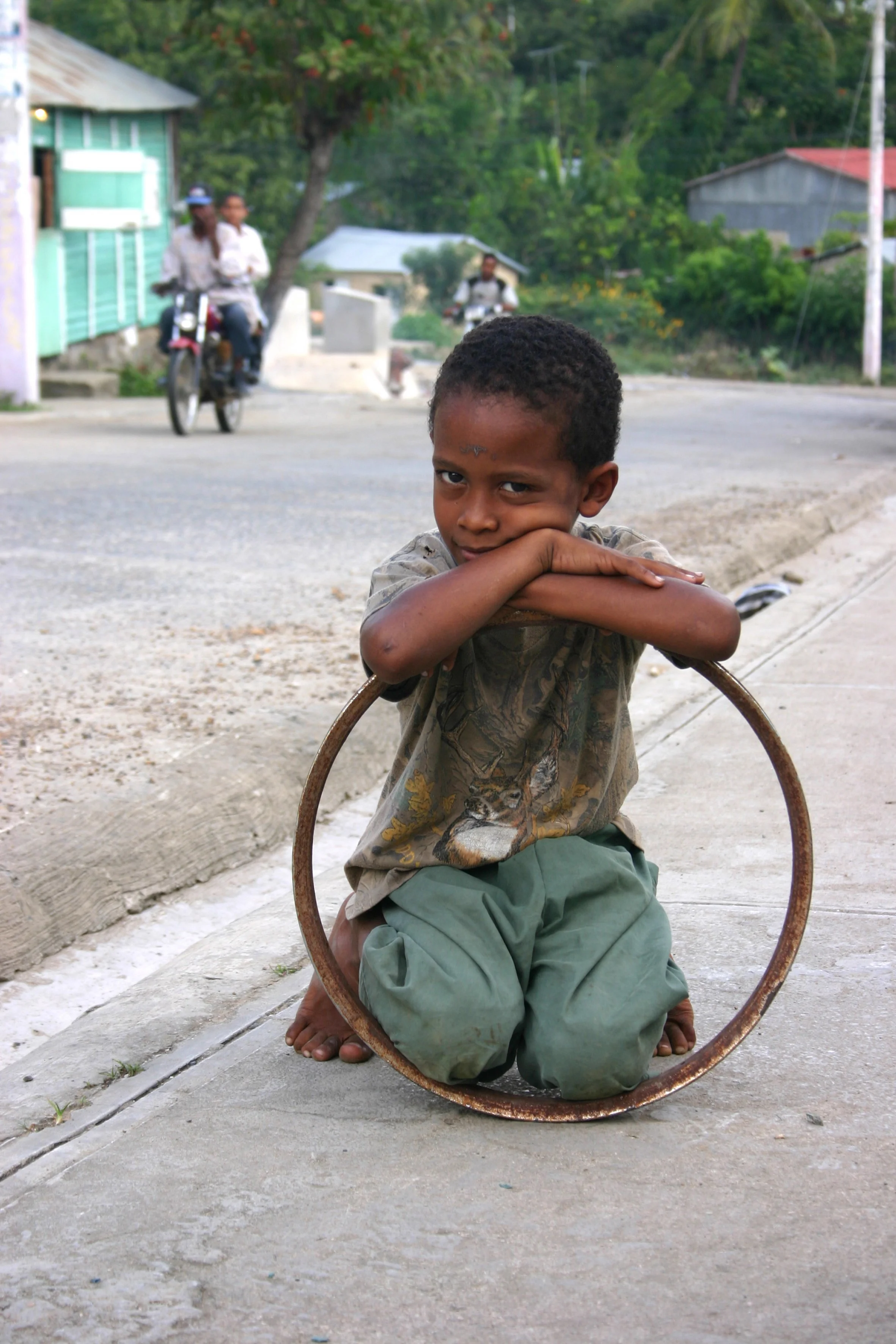Un jeune garçon assis à genoux sur un trottoir, avec un cercle en métal autour de lui, dans un quartier résidentiel en milieu rural. Il porte un t-shirt beige avec un motif et un pantalon vert, et regarde à la caméra avec une expression pensative.