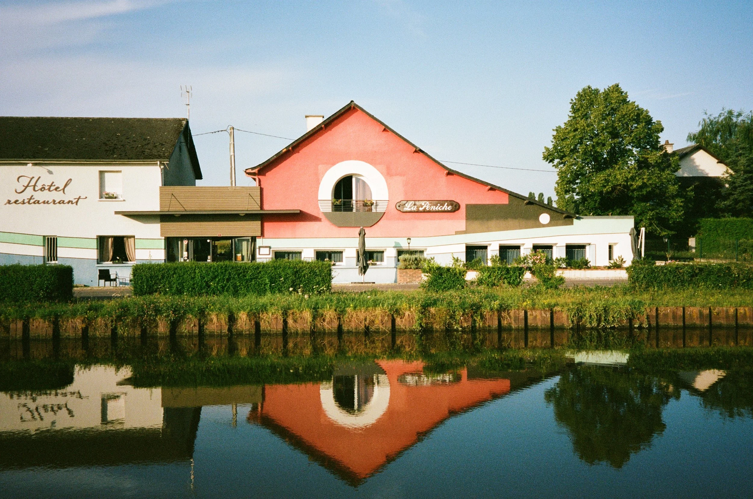 Une maison au bord de l'eau avec un reflet parfait, un bâtiment rose avec un cercle blanc autour d'une fenêtre, et un panneau indiquant 'La Peniche', sous un ciel bleu clair.