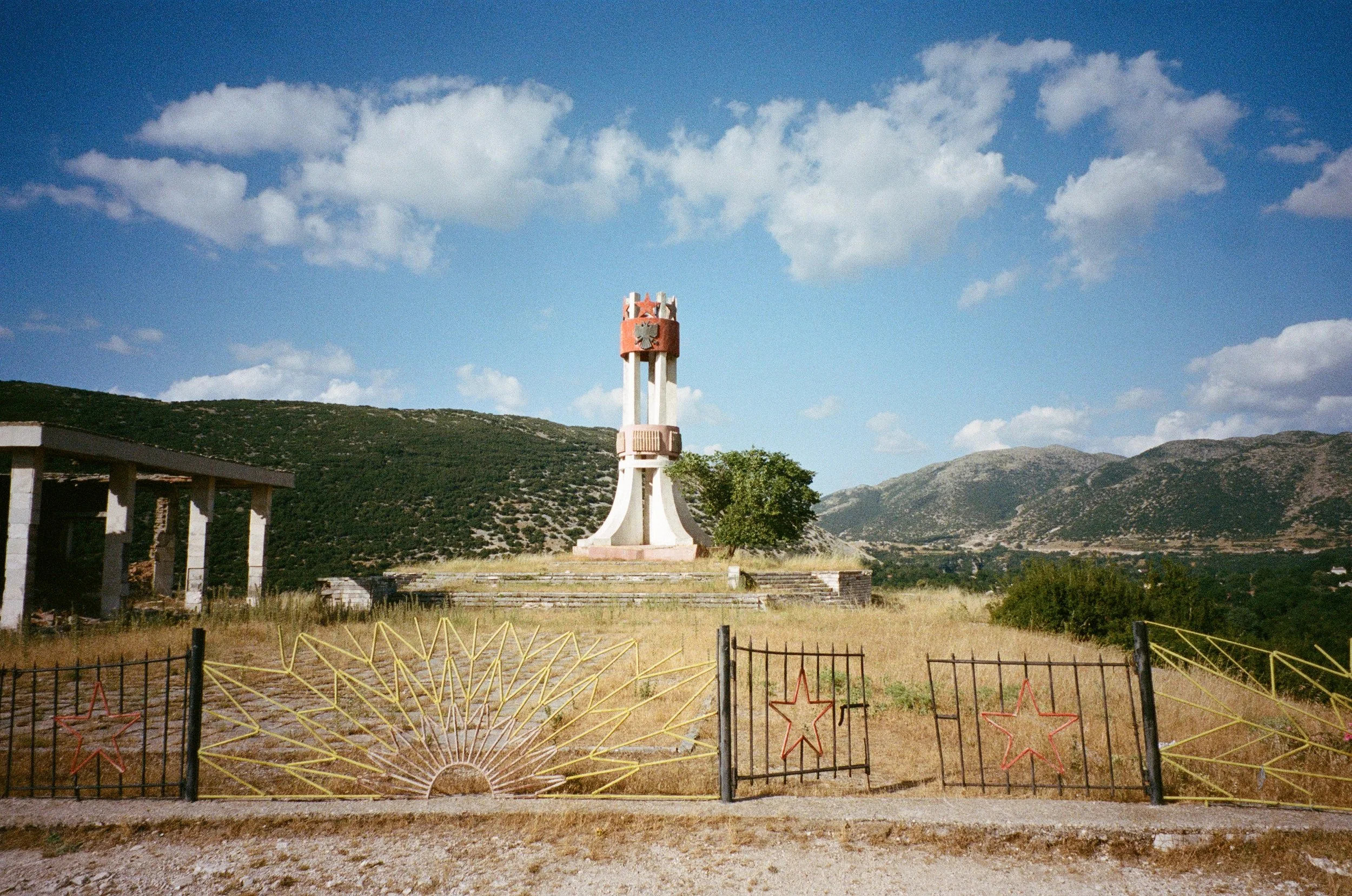 Monument au centre d'une prairie, entouré de jeunes arbres et clôture avec décorations en forme d'étoiles rouges, montagnes en arrière-plan, ciel bleu avec nuages