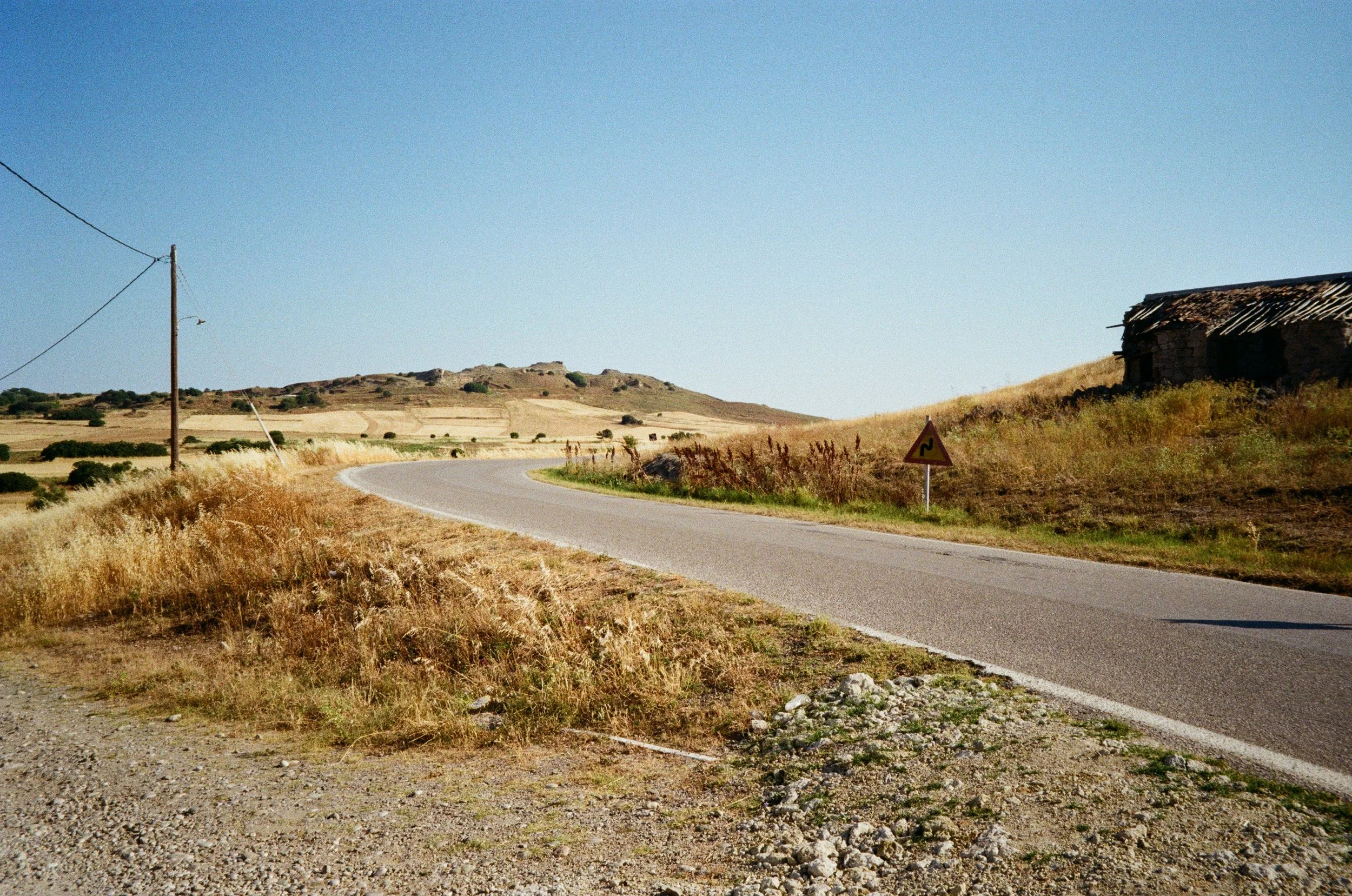 Route de campagne sinueuse avec un panneau de signalisation en haut à droite, terrain herbeux avec des buissons, ancien bâtiment en ruines, terrain en pente douce et un ciel clair.