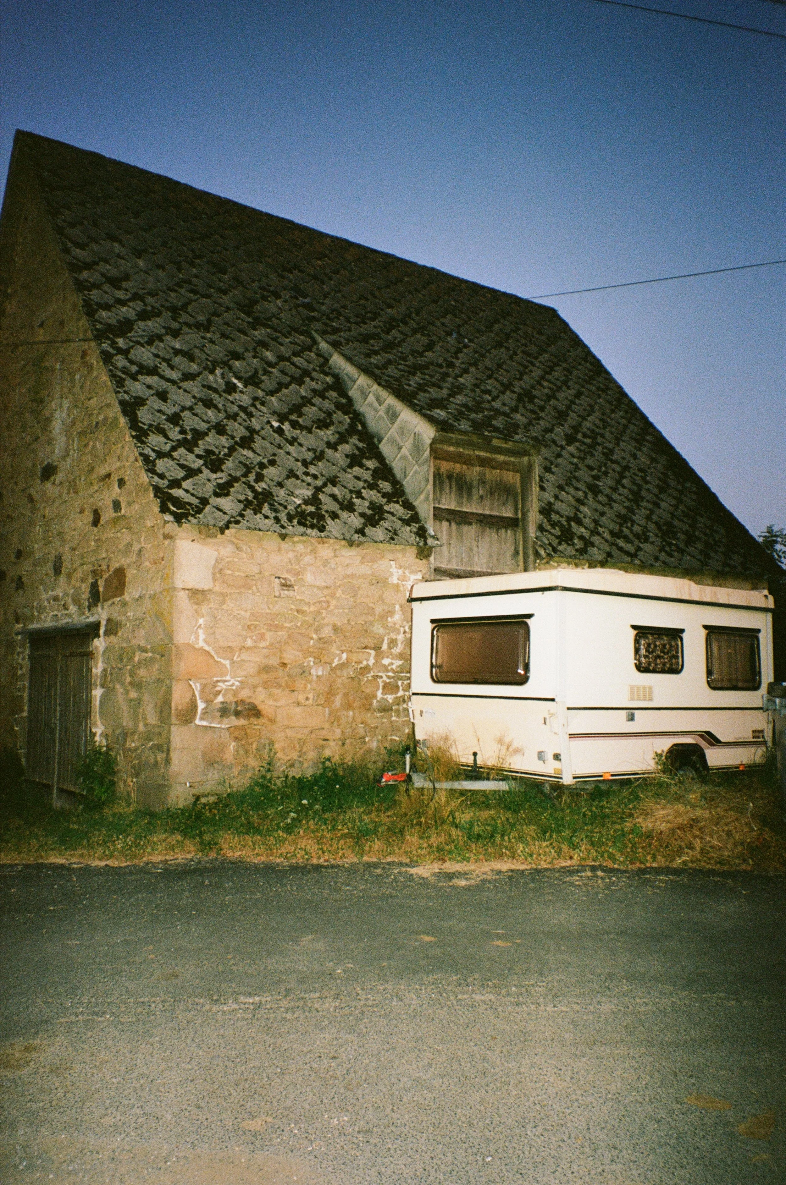 Une caravane blanche stationnée à côté d'une vieille maison en pierre avec un grand toit en ardoise, sous un ciel crépusculaire.