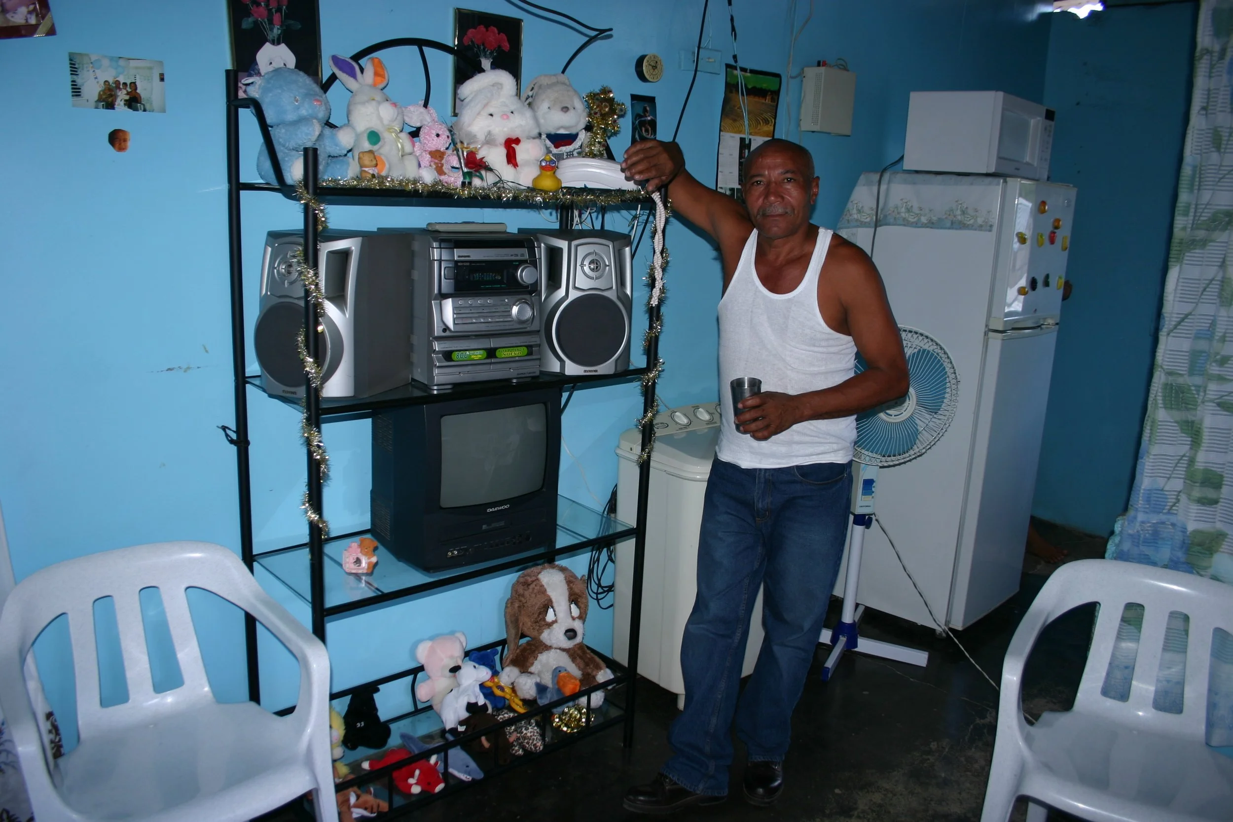 Un homme debout dans une pièce aux murs bleus, tenant une tasse en métal et portant un débardeur blanc. Derrière lui, une étagère noire avec plusieurs peluches, une chaîne de Noël, un vieux téléviseur, une chaîne stéréo, et un réfrigérateur. Deux cha