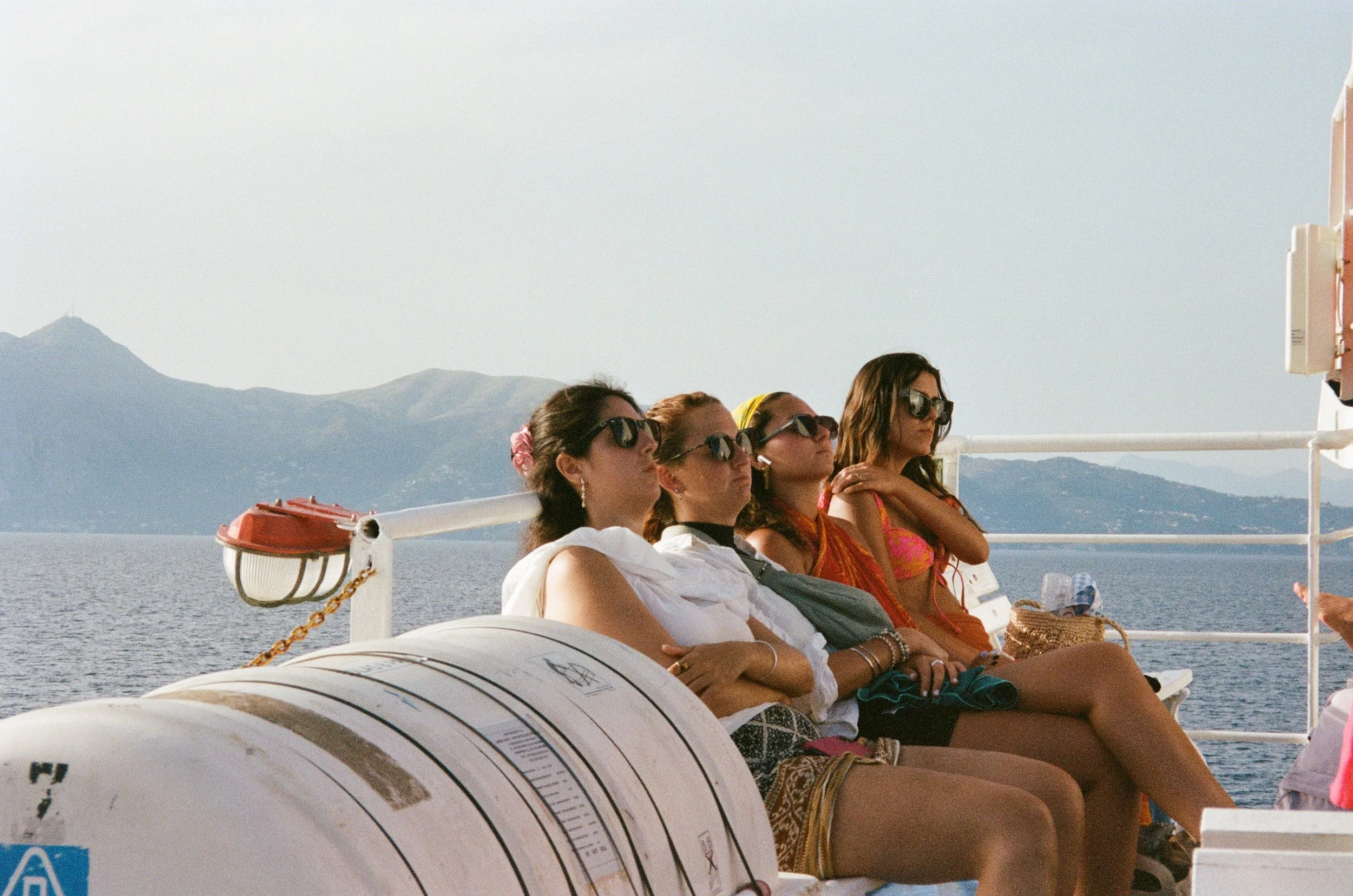Cinq femmes assises sur le pont d'un bateau, portant des lunettes de soleil, regardant droit devant, avec la mer et des montagnes en arrière-plan.