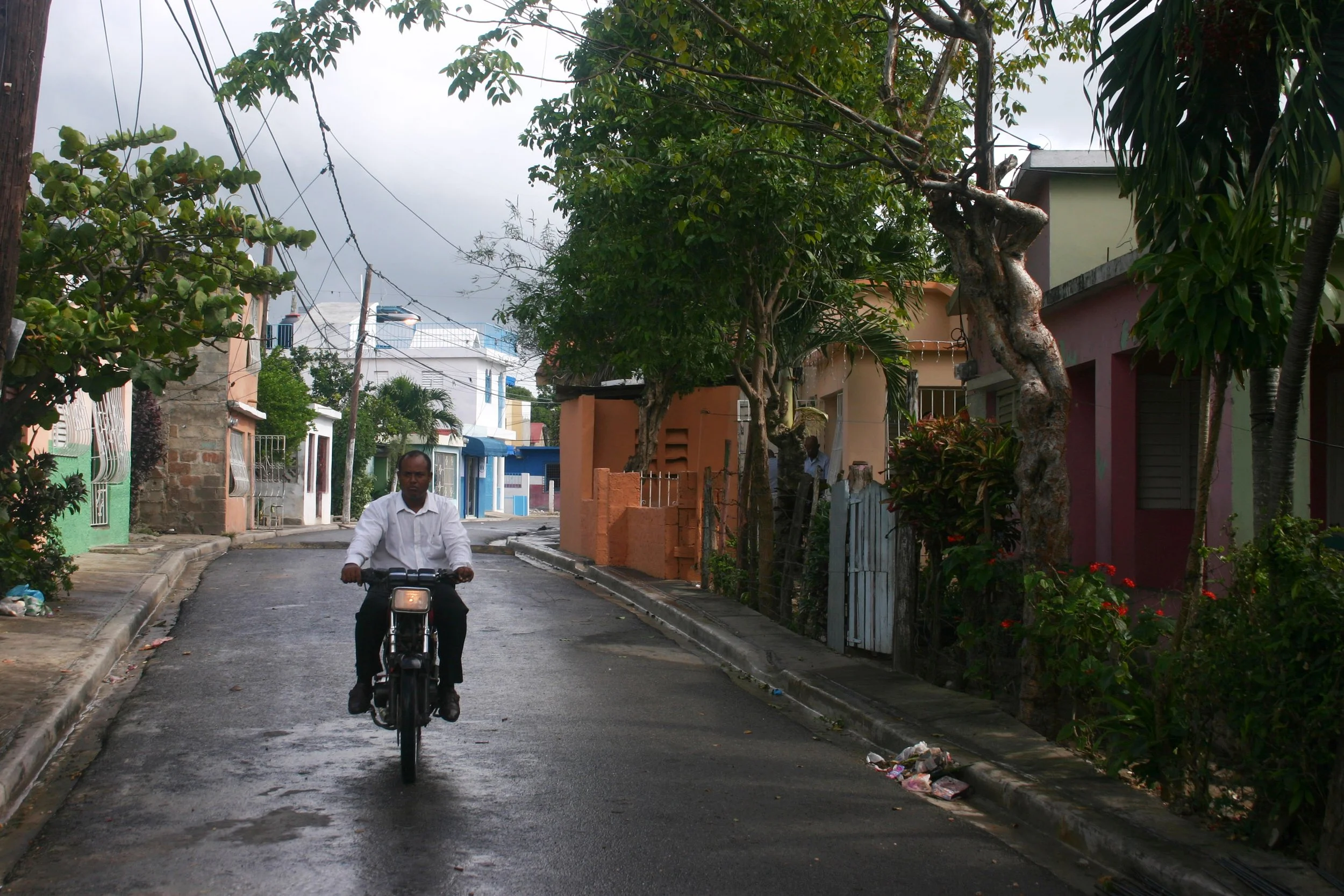 Un homme en chemise blanche pédalant sur une rue pavée dans un quartier résidentiel coloré, avec des maisons et des arbres.