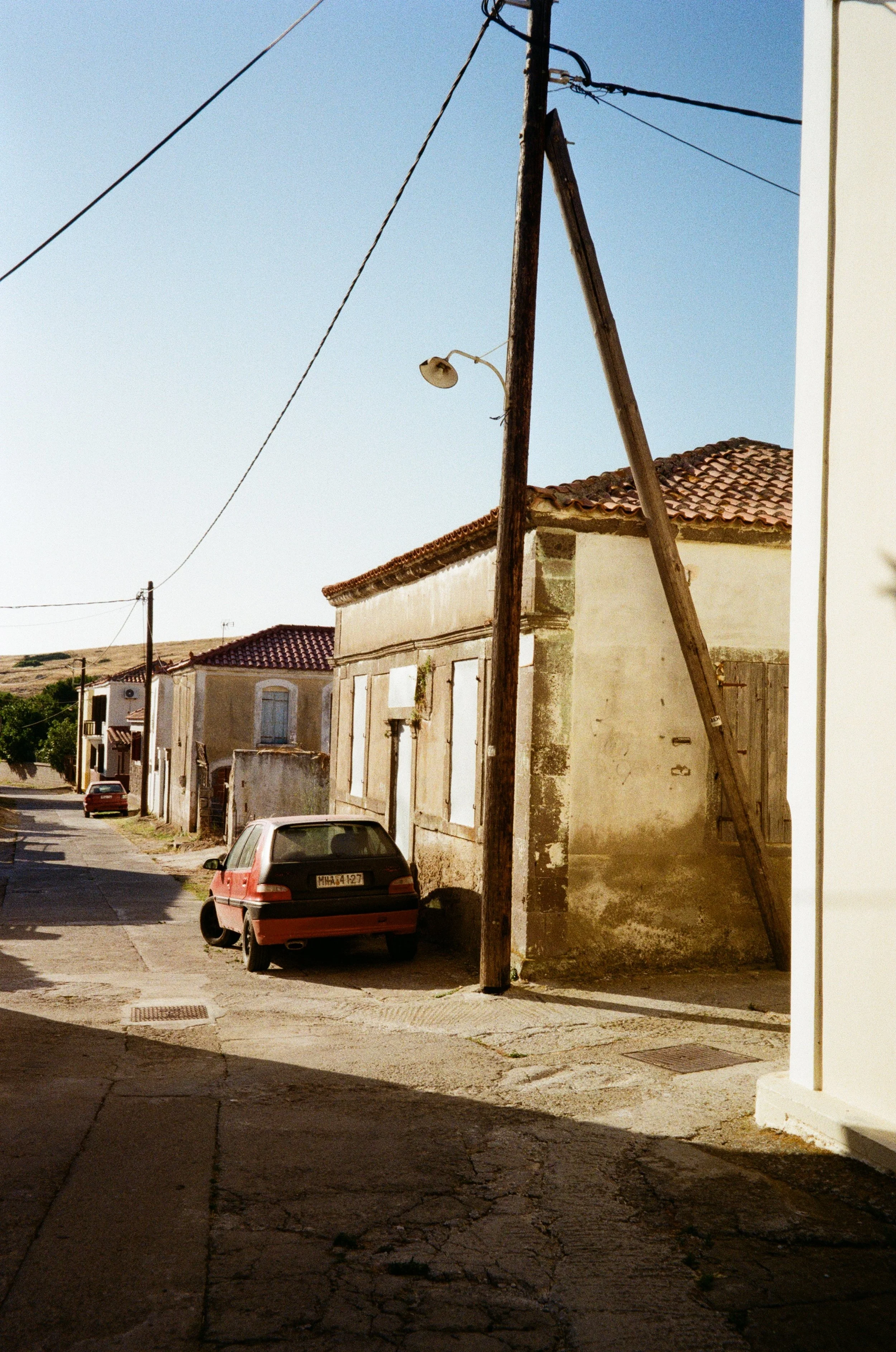 Rue de village avec une voiture rouge garée, maisons avec toits en tuiles, poteau électrique en bois, ciel clair.